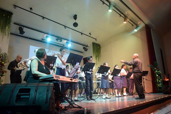 Orquestra de alunos tocando instrumentos sob a regência de um maestro no palco.