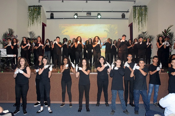 Apresentação de alunos no palco, todos vestidos de preto, fazendo coreografia em grupo.