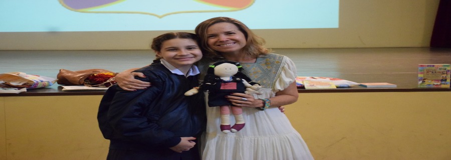 Menina e mulher sorrindo no palco, abraçadas, segurando uma boneca artesanal.