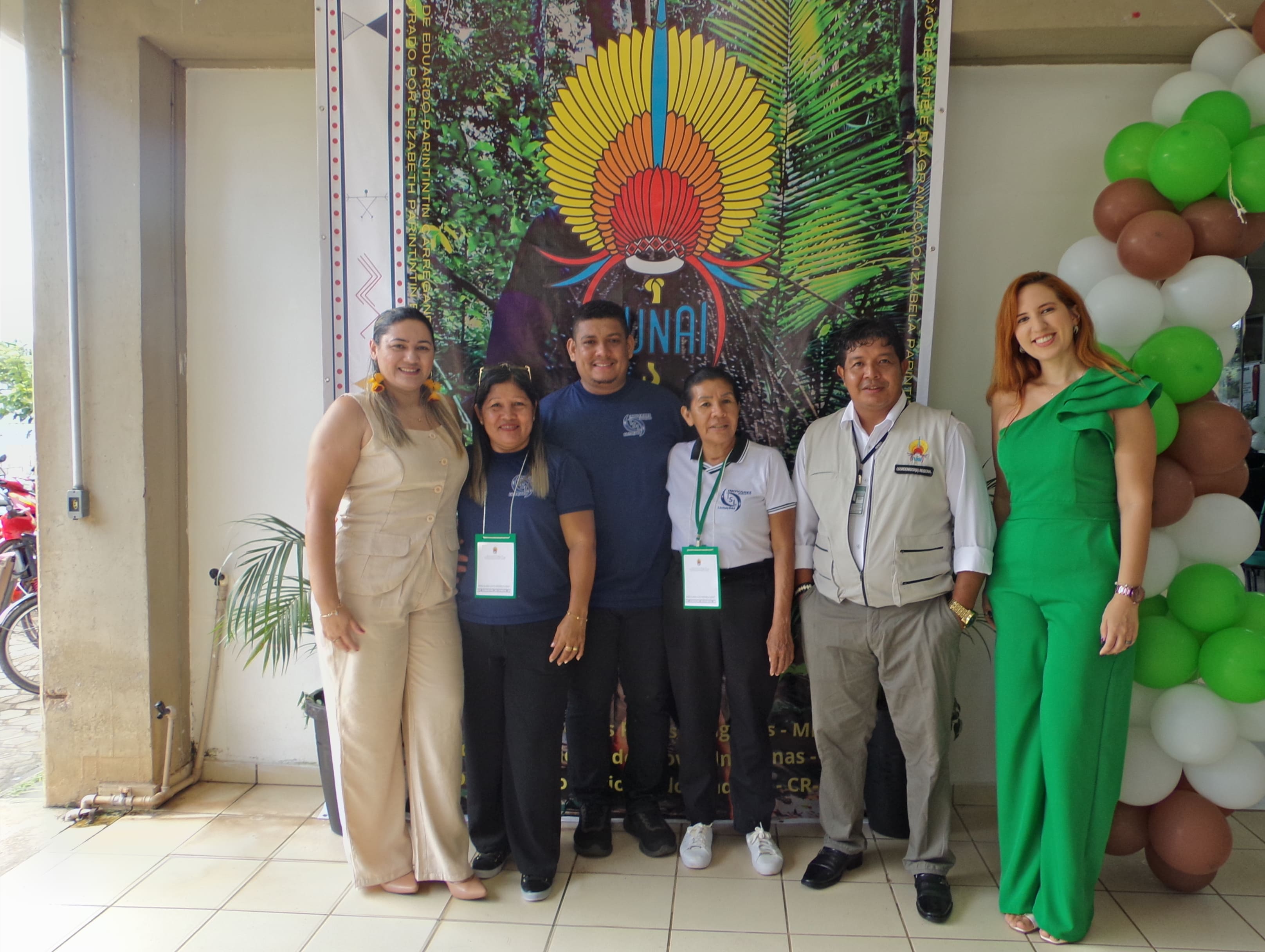 Grupo de seis pessoas posando diante de um banner colorido com arte indígena e balões decorativos