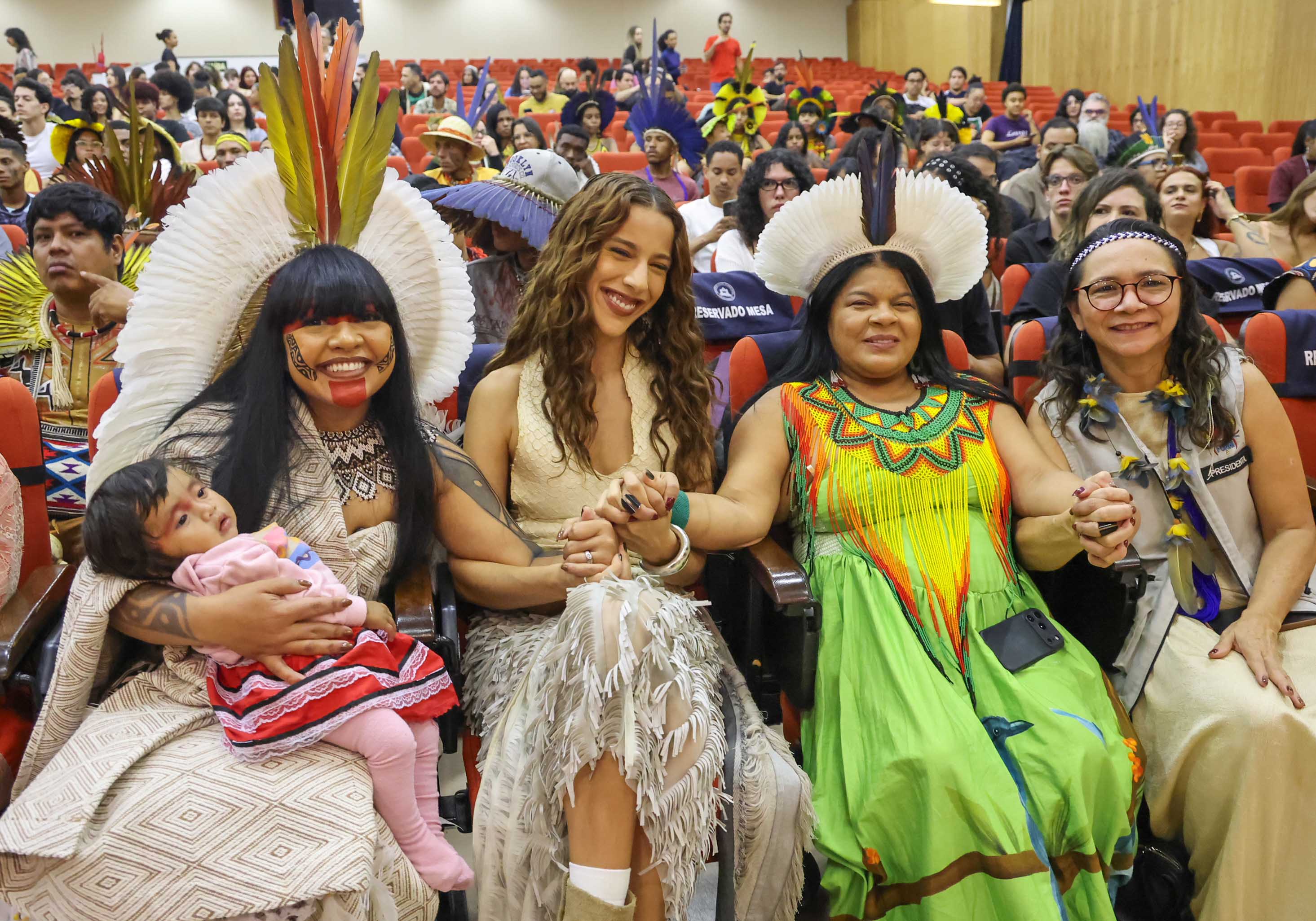 4 mulheres sentadas de mãos dadas com adereços indígenas