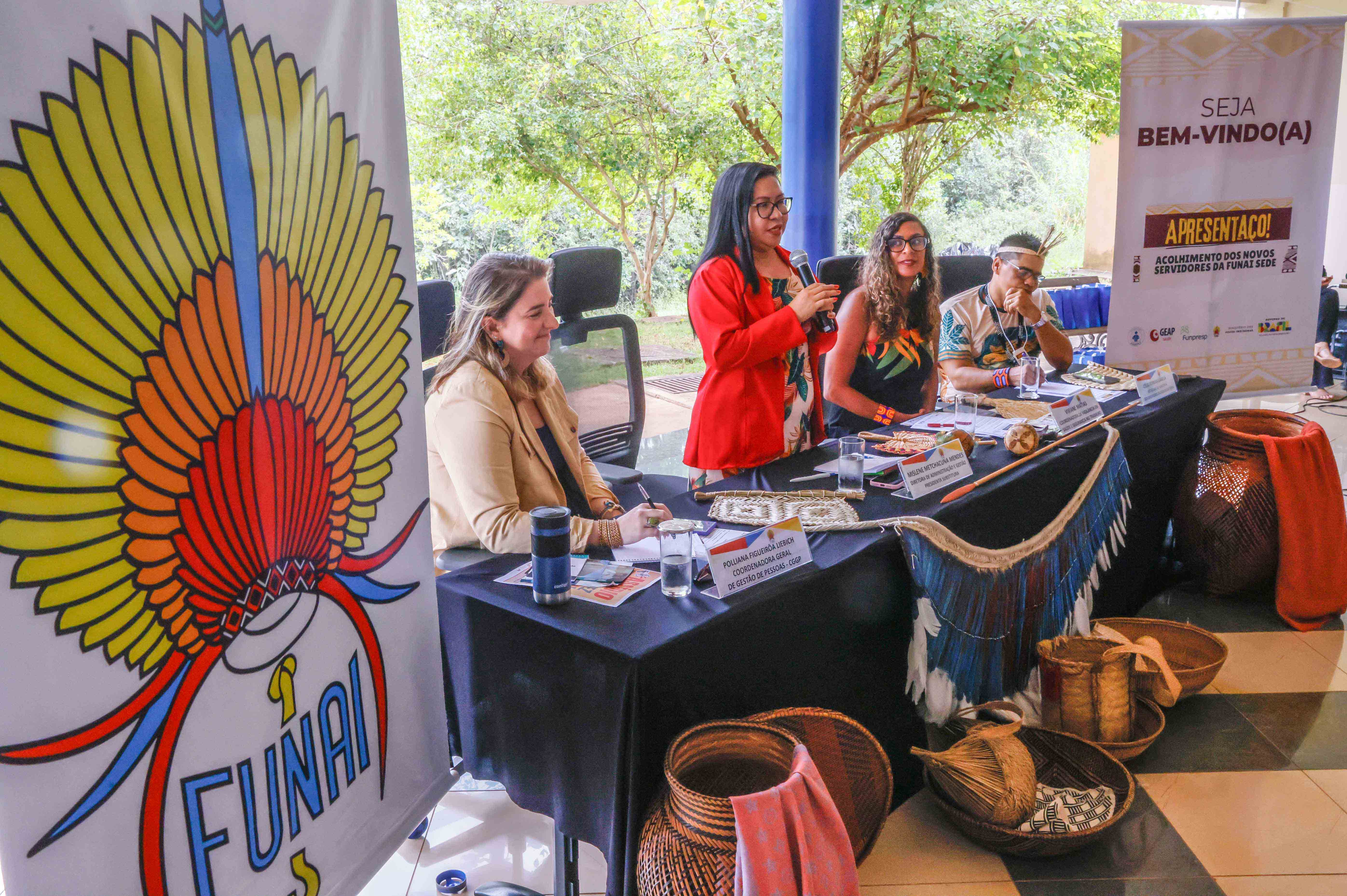 mesa diretora com 4 pessoas, uma mulher em pé falando ao microfone