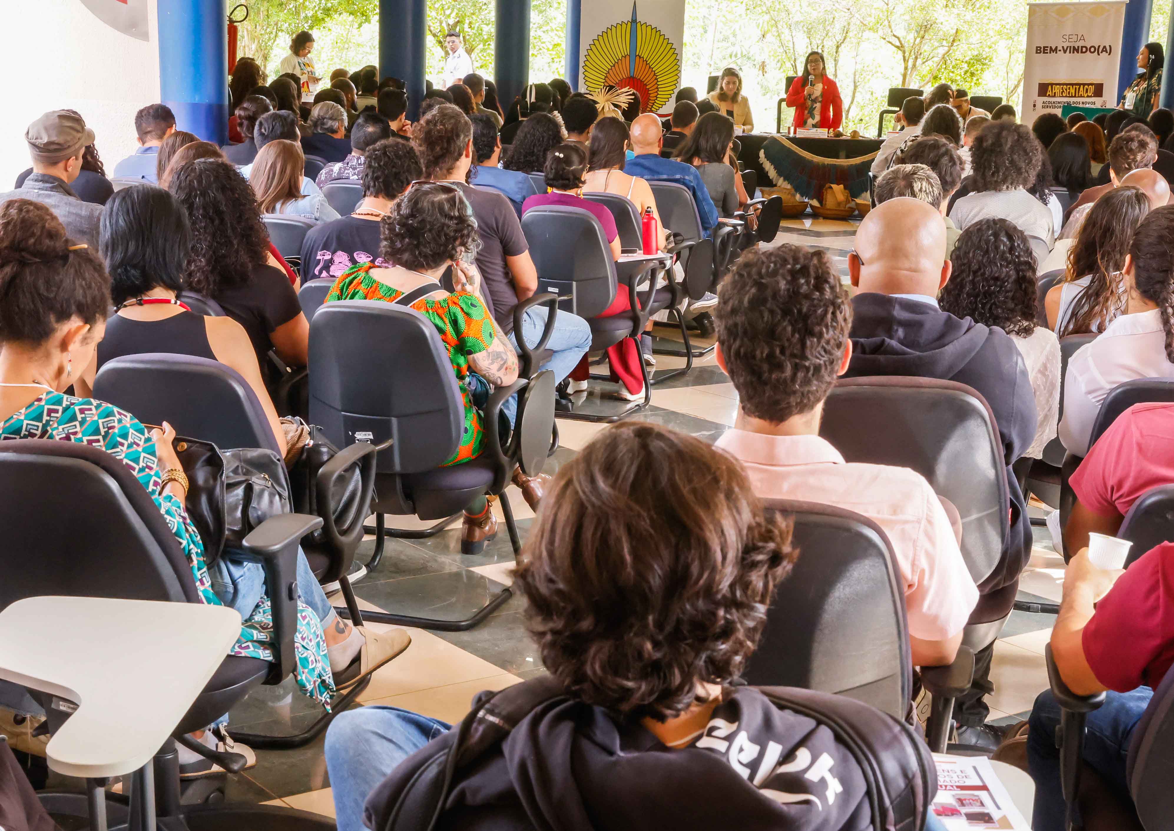 Diversas pessoas sentadas e uma mesa diretora ao fundo com presidenta substituta falando em pé