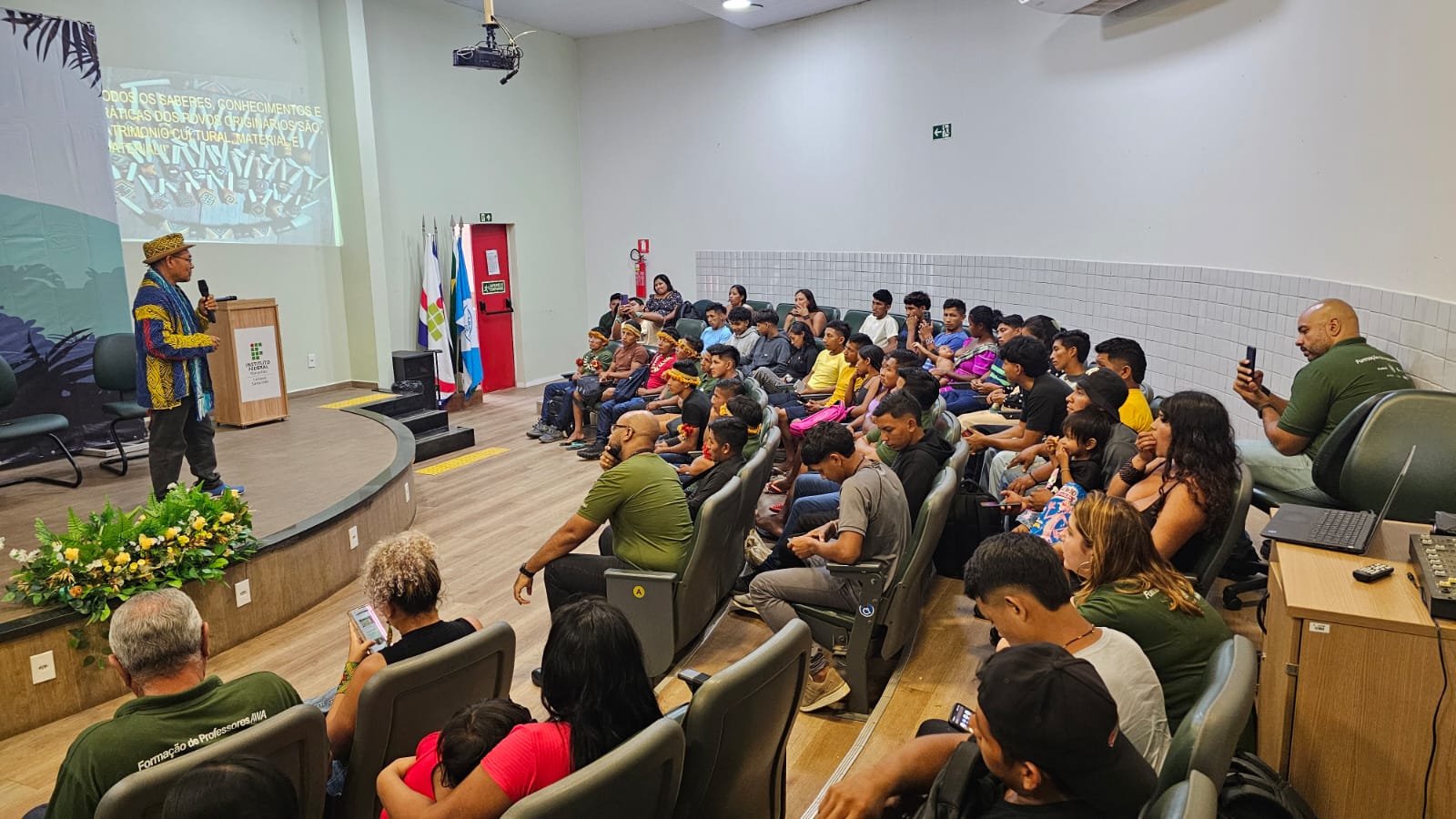 Indígenas sentados em um auditório redondo, com um professor indígena fazendo a aula inaugural e um slide na parede