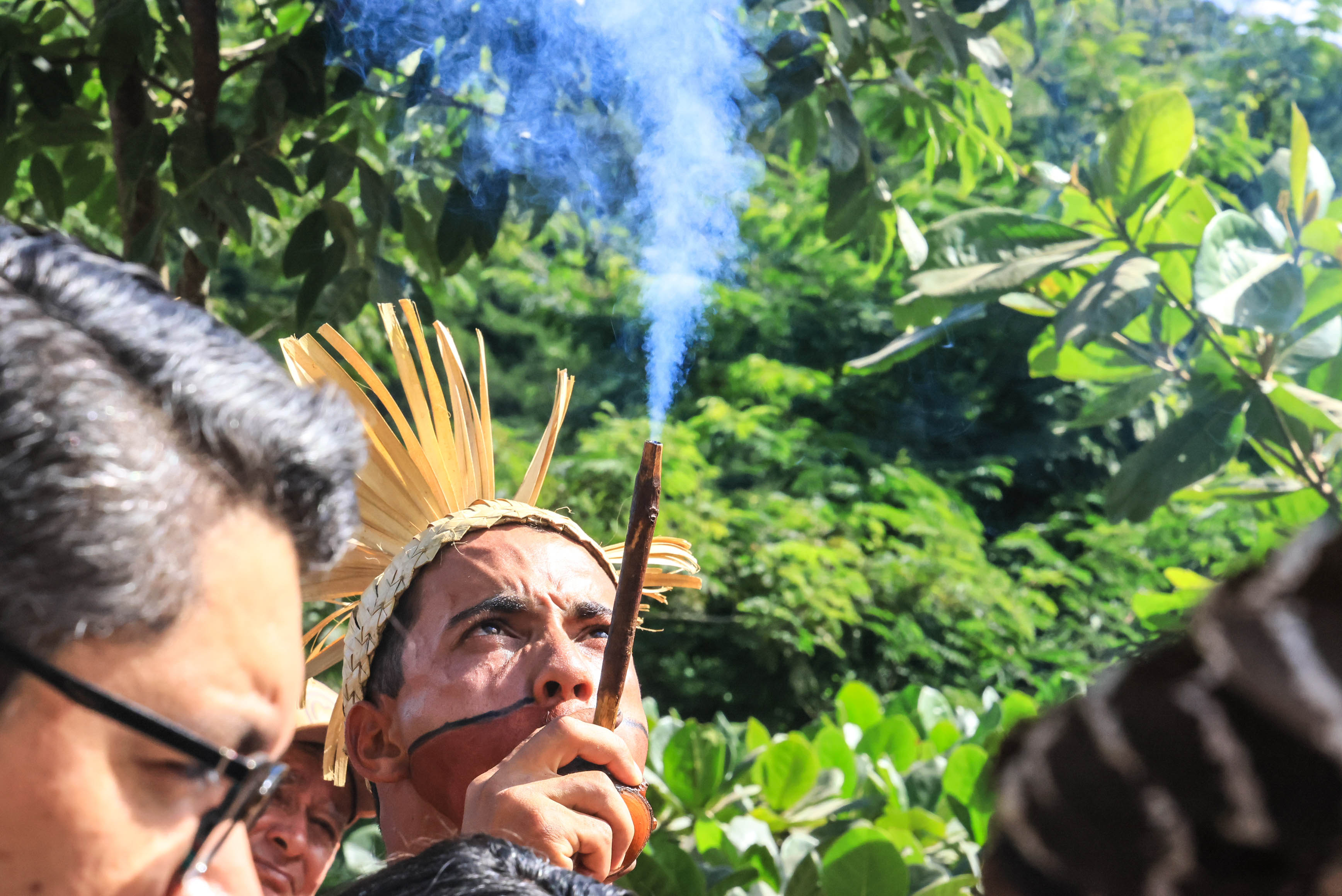 Homem indígena do povo Xukuru solta fumaça por um cachimbo. Usa acessórios tradicionais