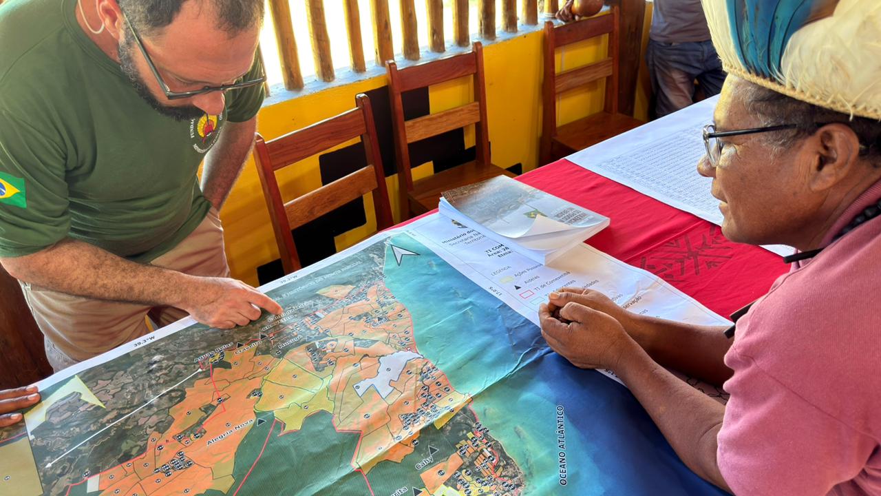 Servidor aponta para localidade em um mapa colocado sobre uma mesa enquanto homem indígena observa