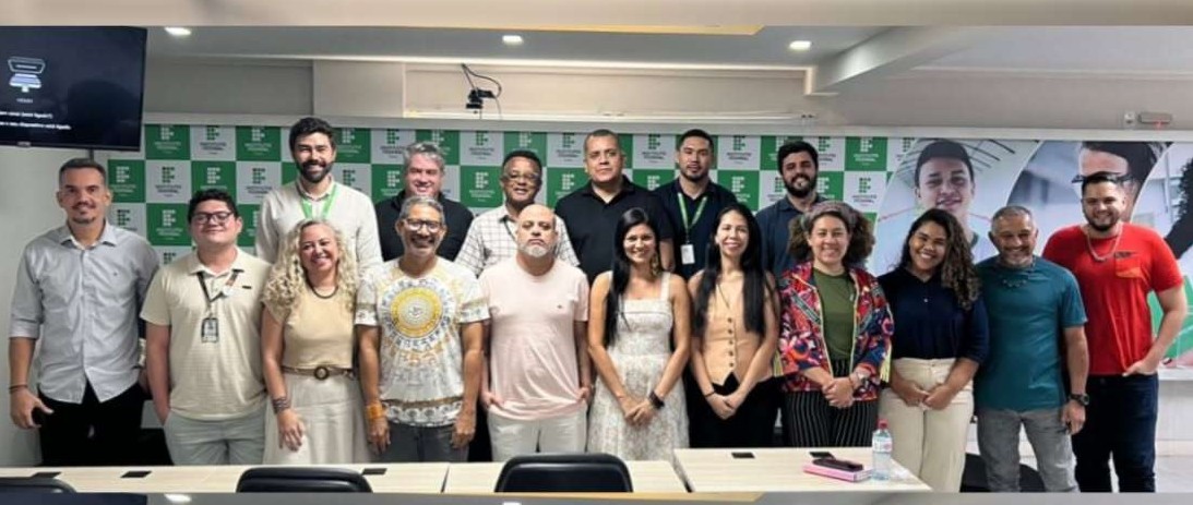 Descrição alt: Grupo de servidores posa para foto em uma sala de reunião da Funai, em frente a um painel institucional verde e branco. Ao todo, 16 pessoas aparecem em pé, organizadas em duas fileiras, sorrindo para a câmera. O ambiente possui mesas, cadeiras e iluminação interna, em um momento de integração entre os participantes.