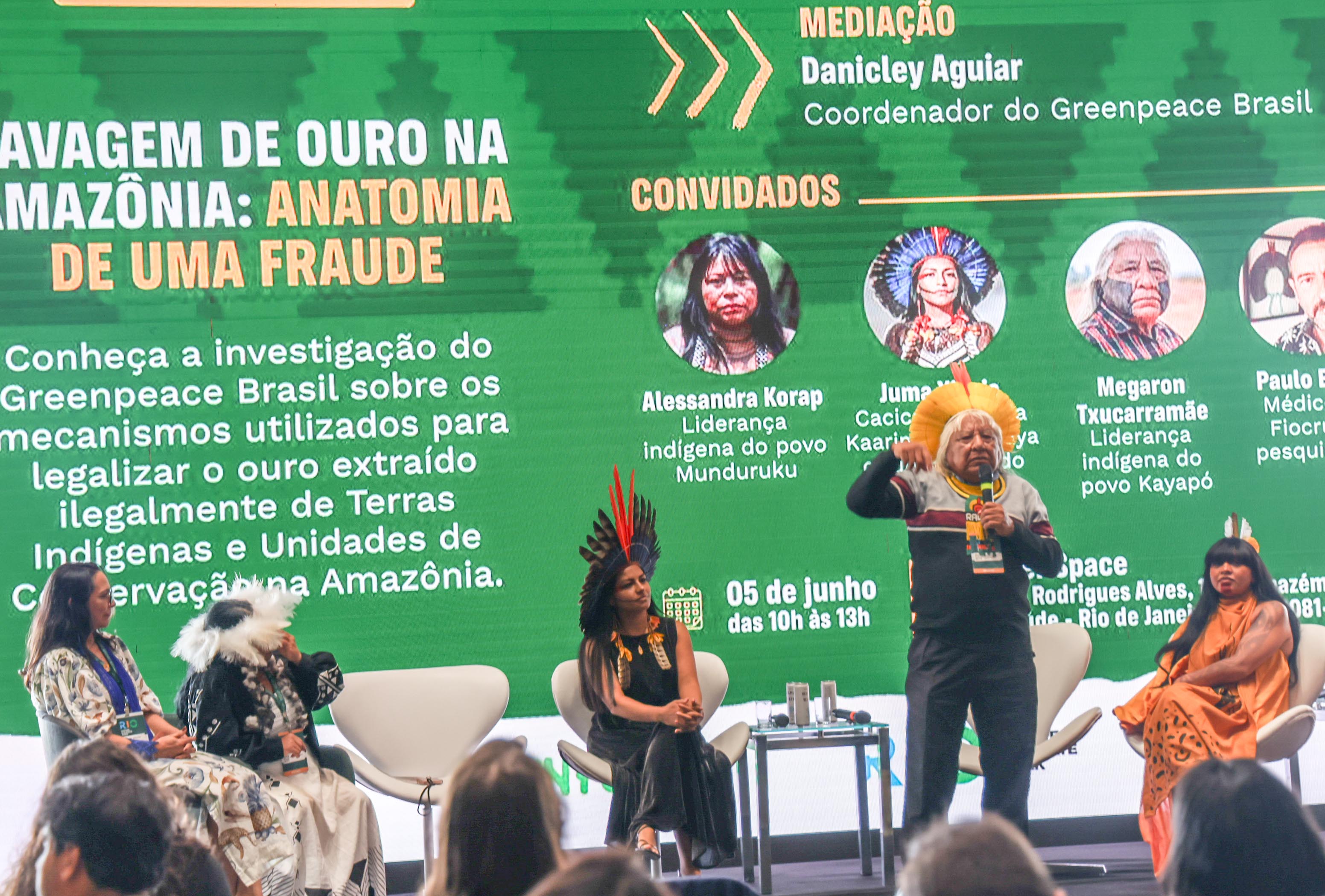 Cacique Raoni falando em pé no palco e 4 mulheres sentadas em cadeiras no palco