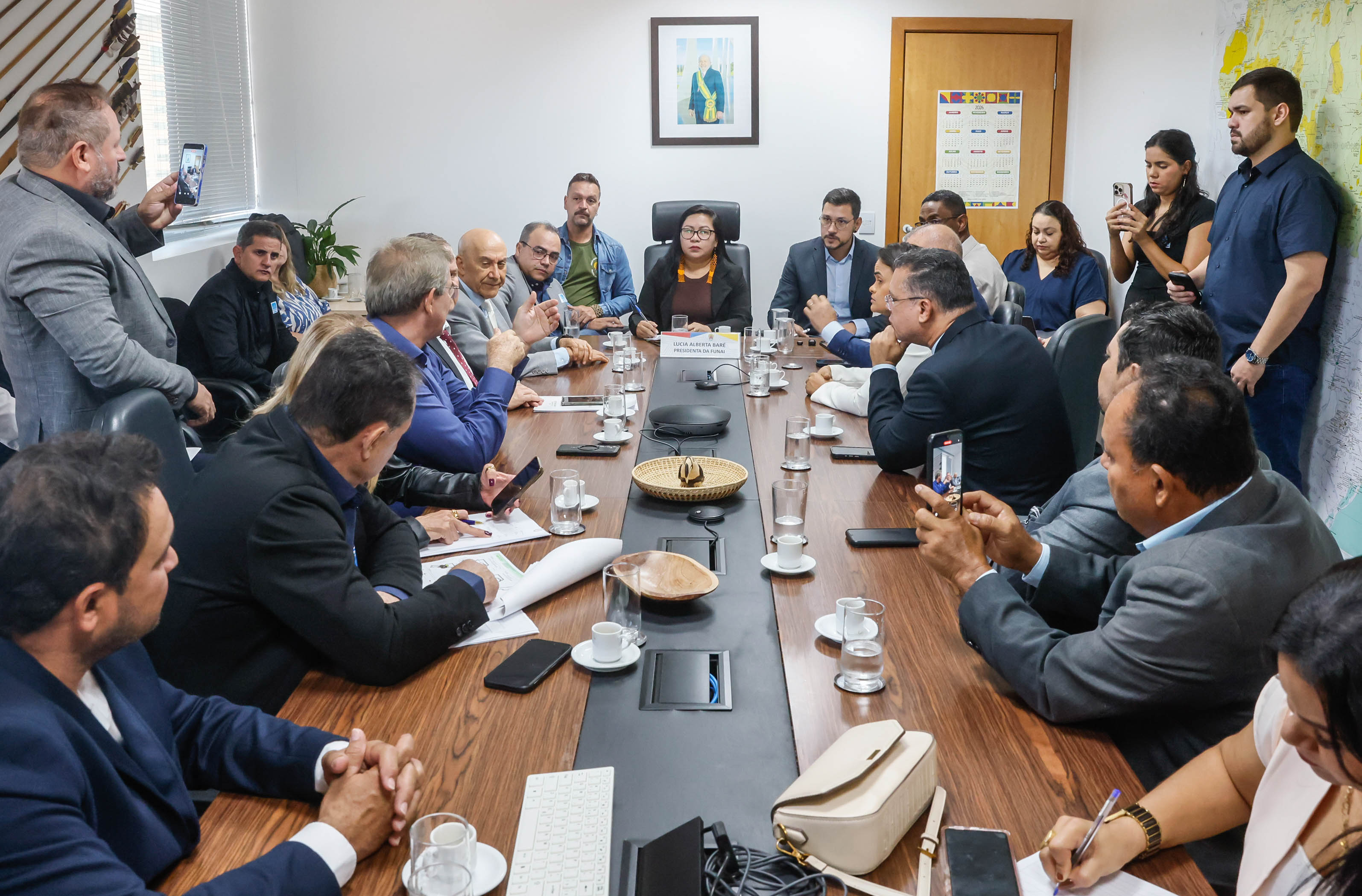 Muitas pessoas em redor de uma mesa em uma reunião, algumas pessoas em pé registrando