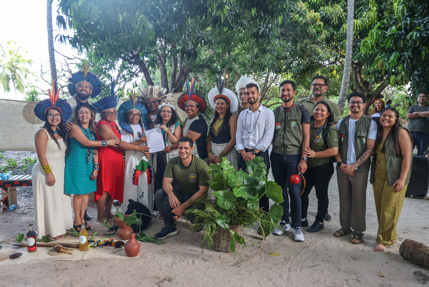 Grupo posa ao ar livre com cocares e documentos, ao lado de objetos e plantas no chão.