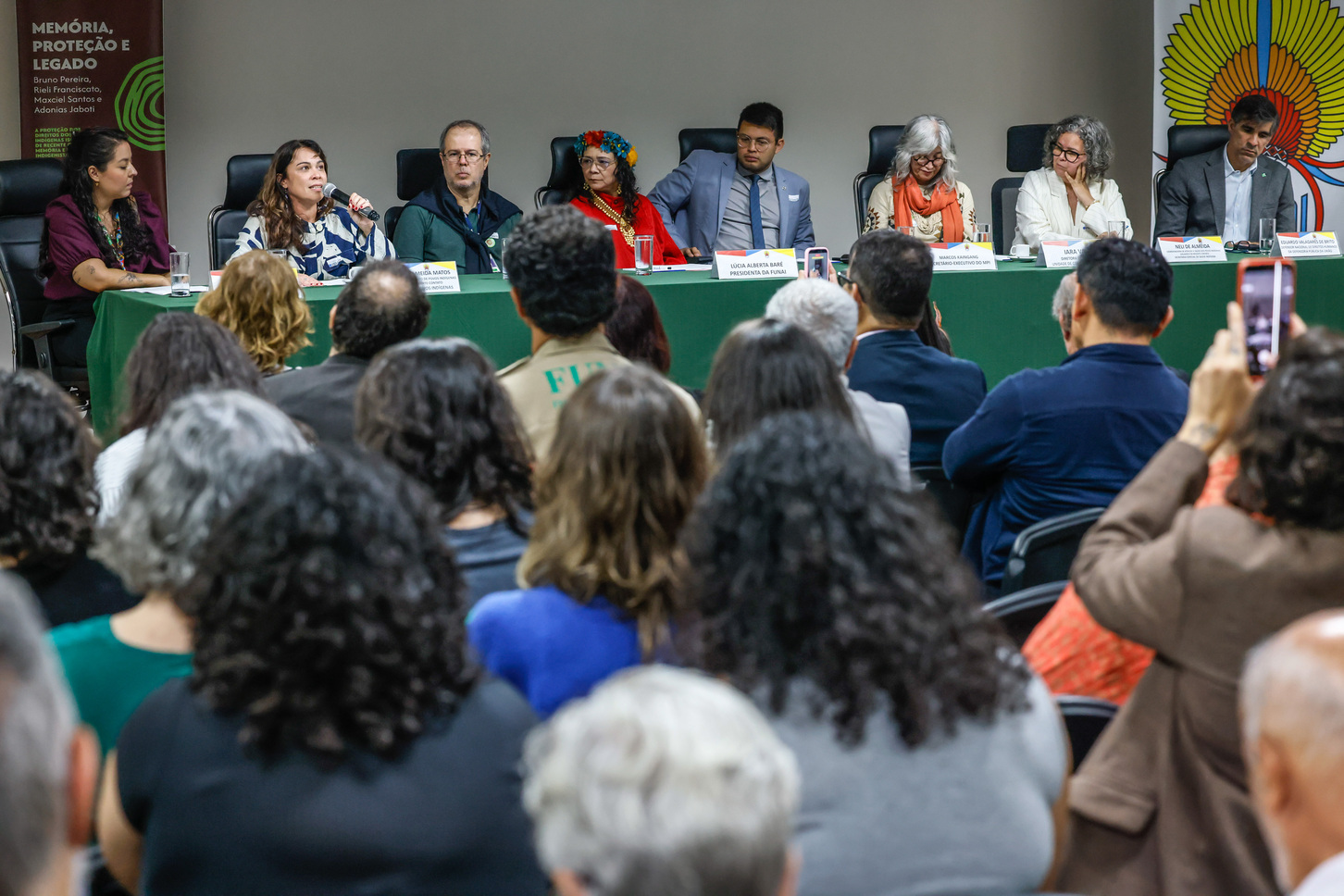Foto de mesa-redonda com sete palestrantes e público em evento institucional.