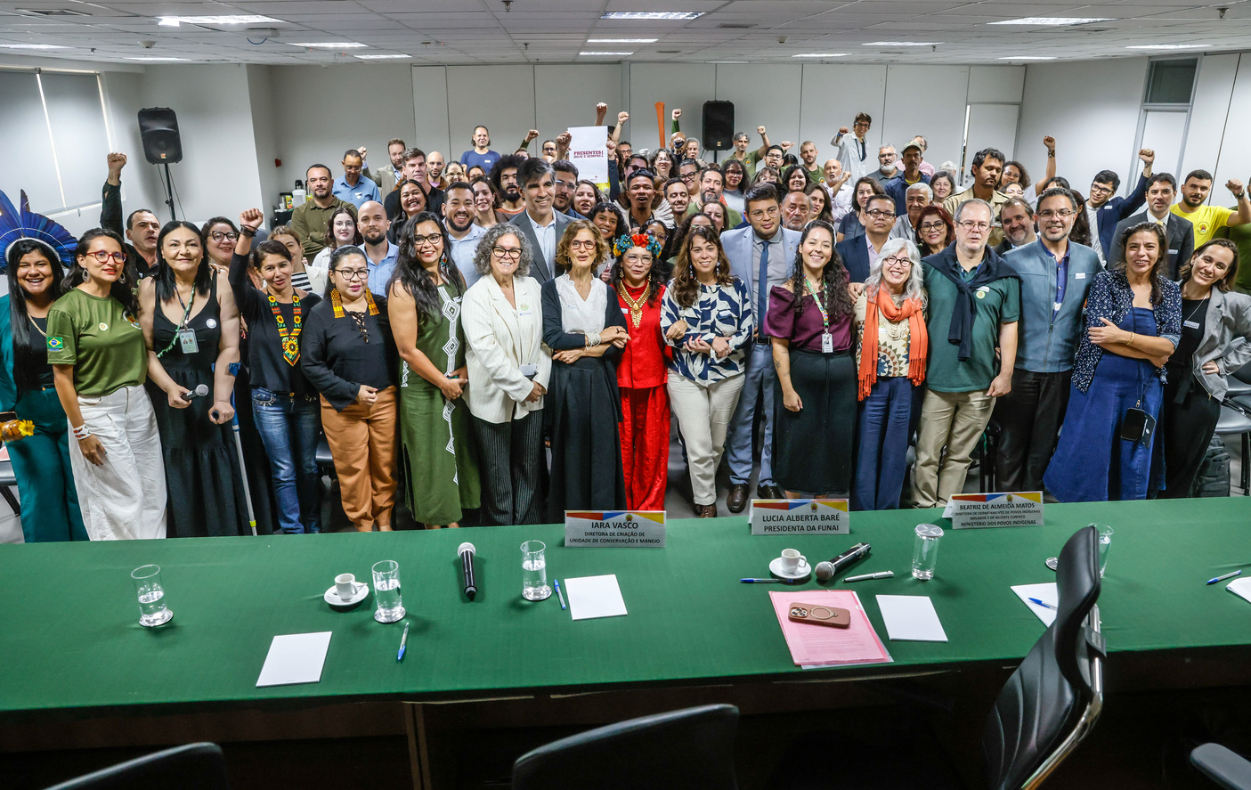 Foto de grupo diverso sorrindo em evento oficial com mesa de autoridades à frente.