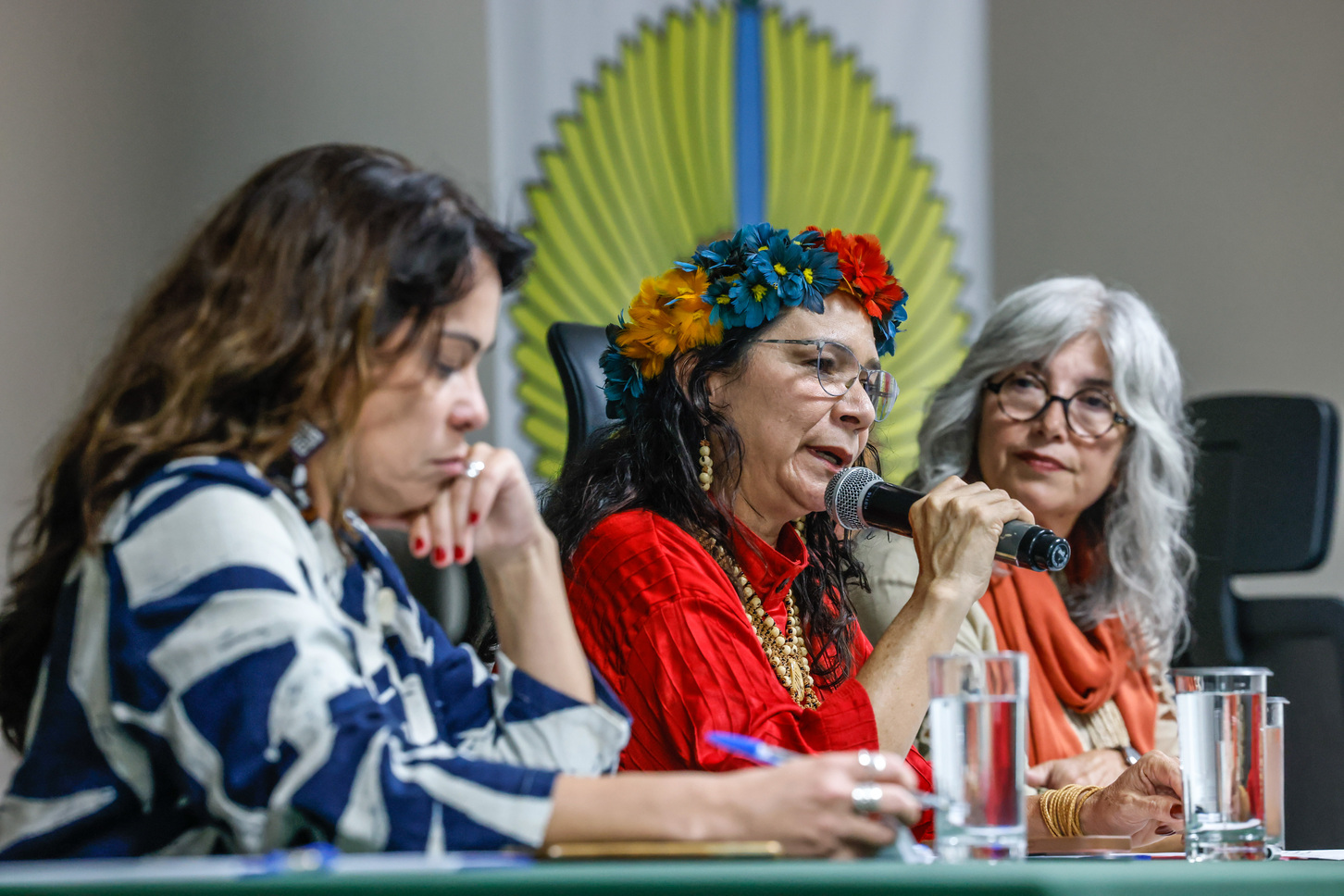 Foto de três mulheres em mesa de debate, uma delas fala ao microfone.