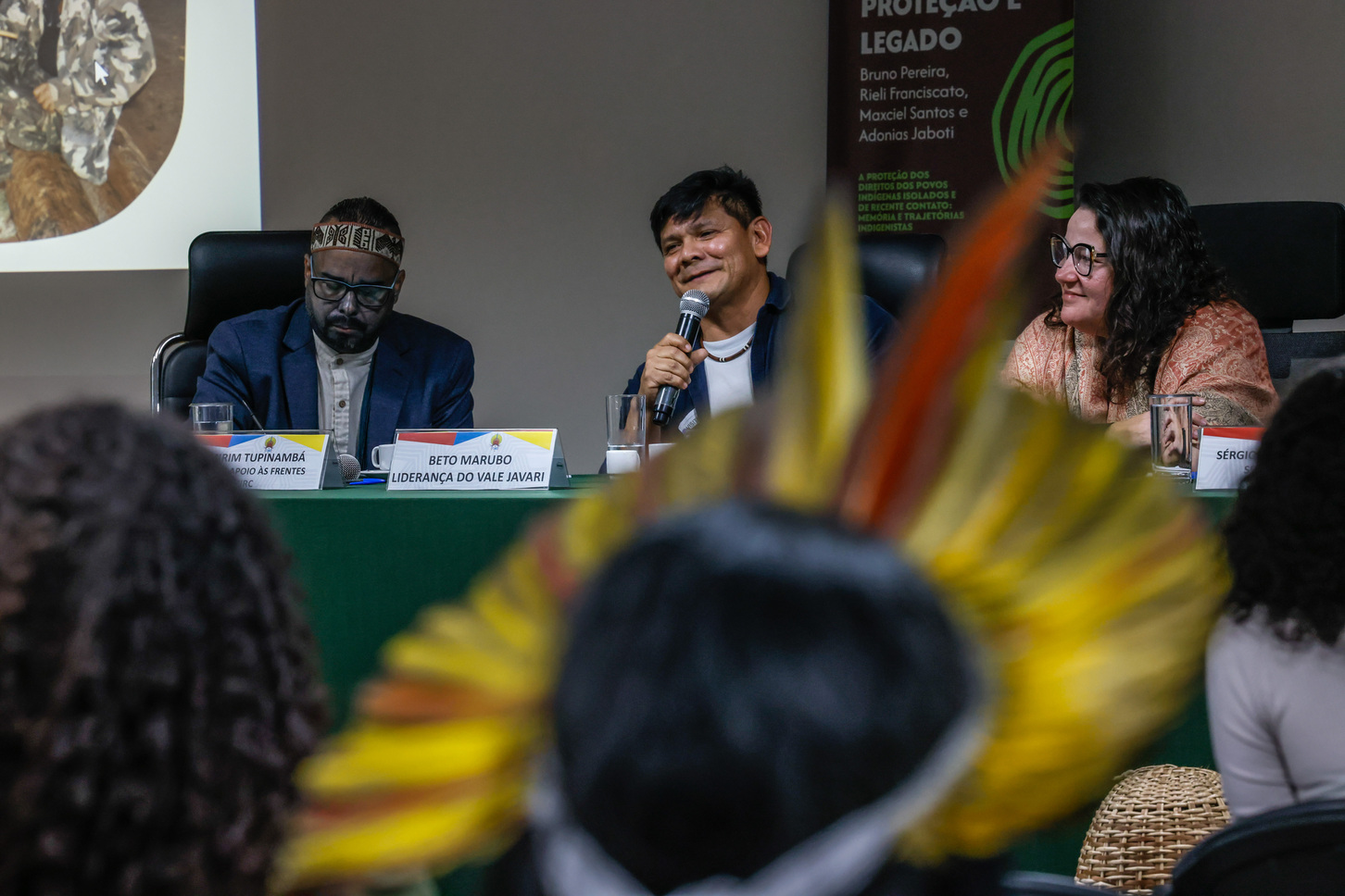Foto de mesa de debate com três participantes, um deles fala ao microfone sorrindo.