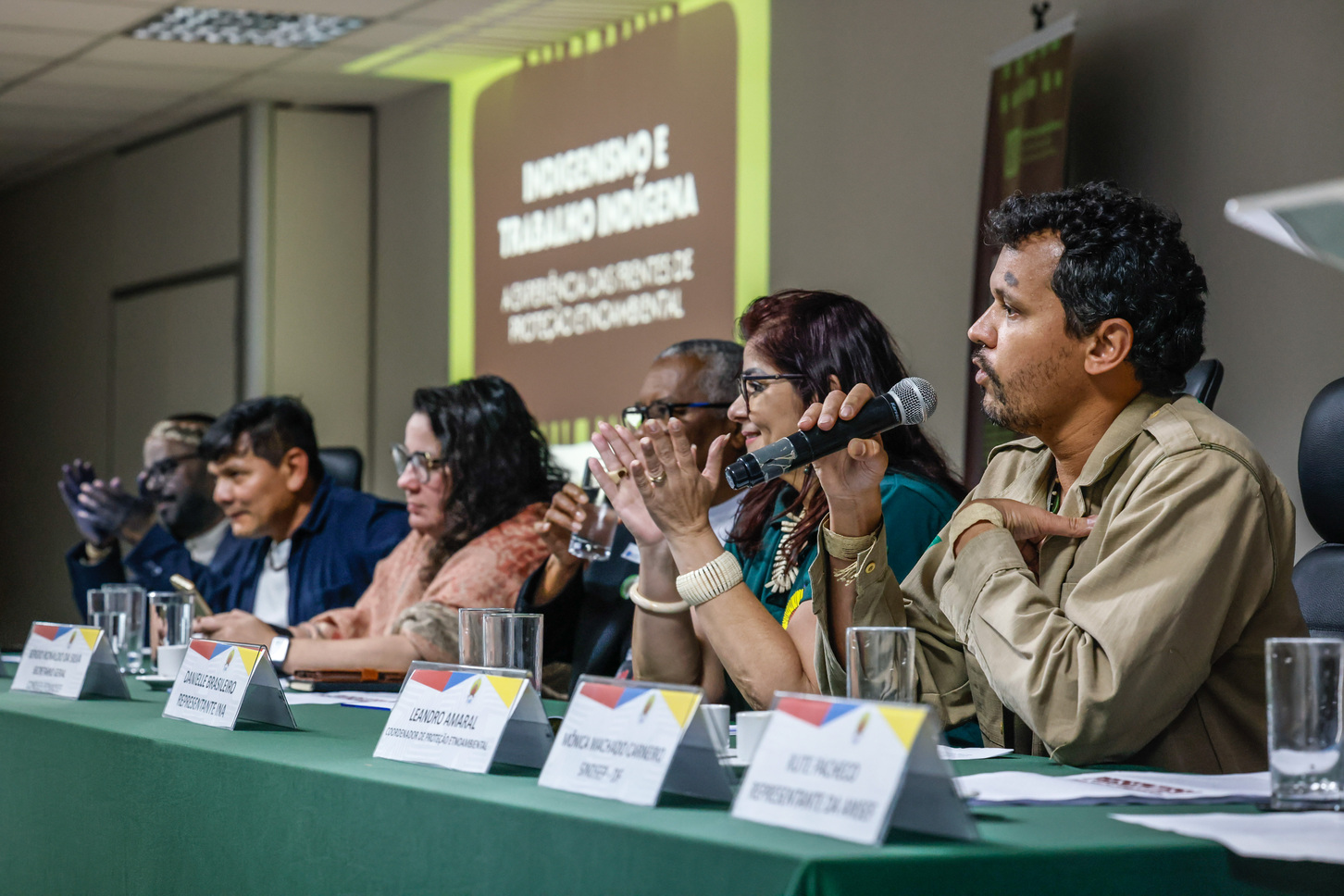 Foto de mesa de debate com seis participantes, um deles fala ao microfone.