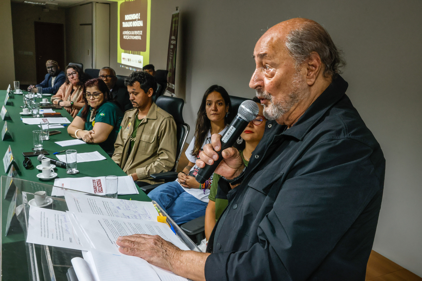 Foto de homem falando ao microfone em mesa de debate com participantes atentos.