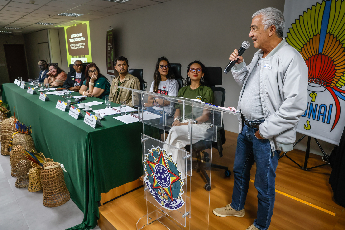 Foto de homem falando ao microfone diante de mesa com sete participantes em evento oficial.