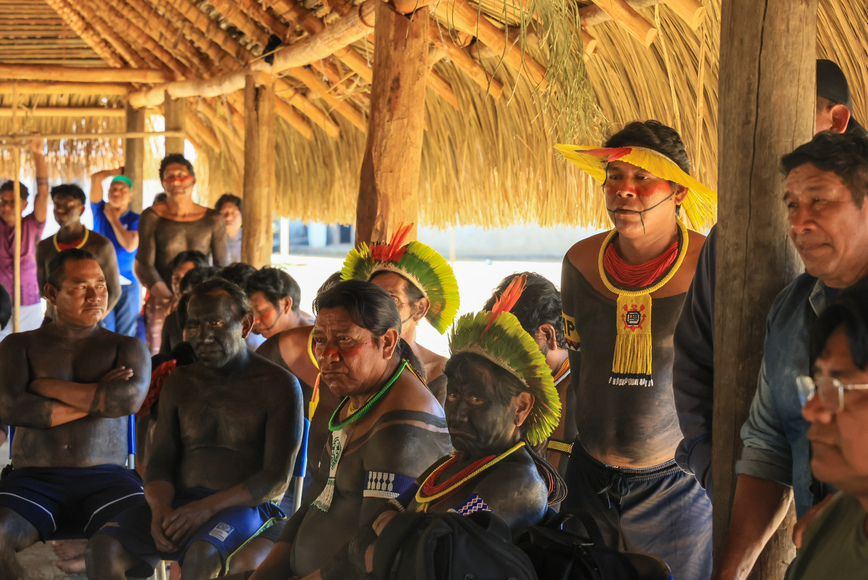 Homens indígenas com pintura corporal e cocares sentados, atentos a uma fala.