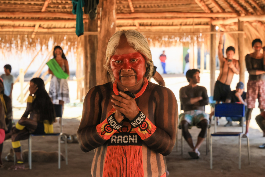 Mulher indígena com pintura facial vermelha e pulseiras "Raoni" posa de frente.