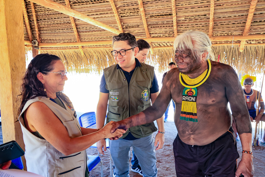 Mulher aperta a mão de cacique indígena; homem de colete observa sorrindo.