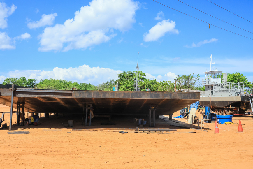 Estrutura metálica de balsa em construção sob céu azul, perto de embarcação.