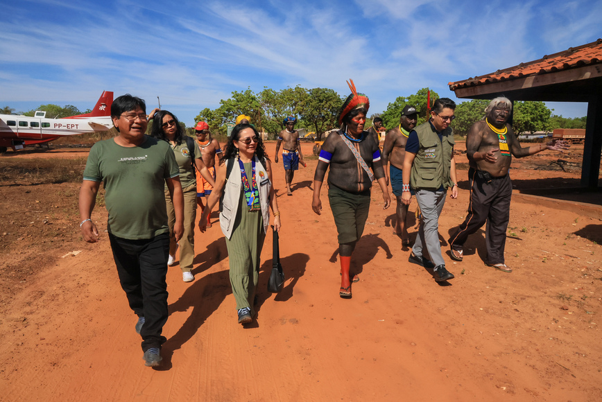 Grupo caminha por estrada de terra; avião pequeno visível atrás.