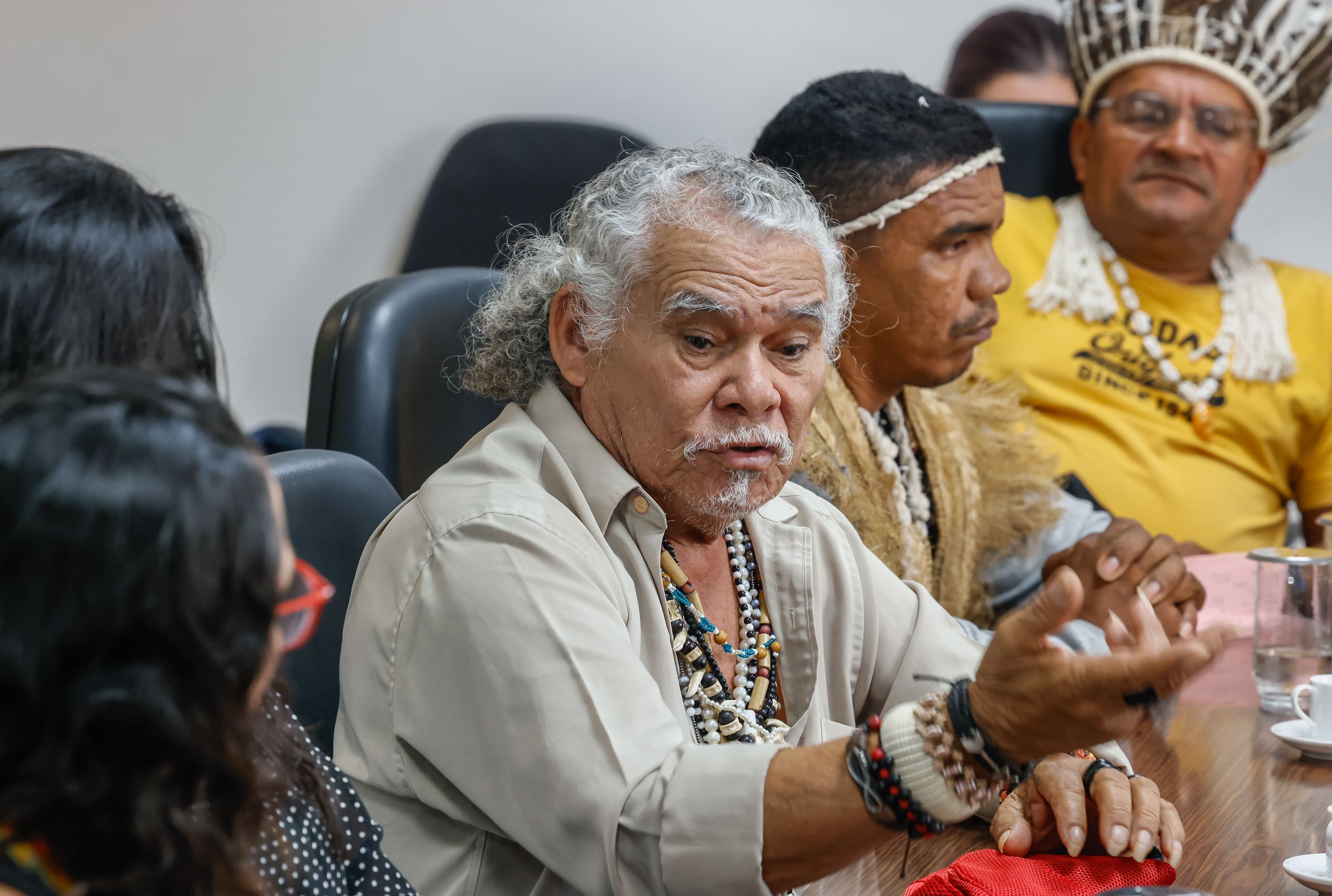 Ancião indígena de cabelos e bigode brancos, vestindo colares artesanais, fala gesticulando.