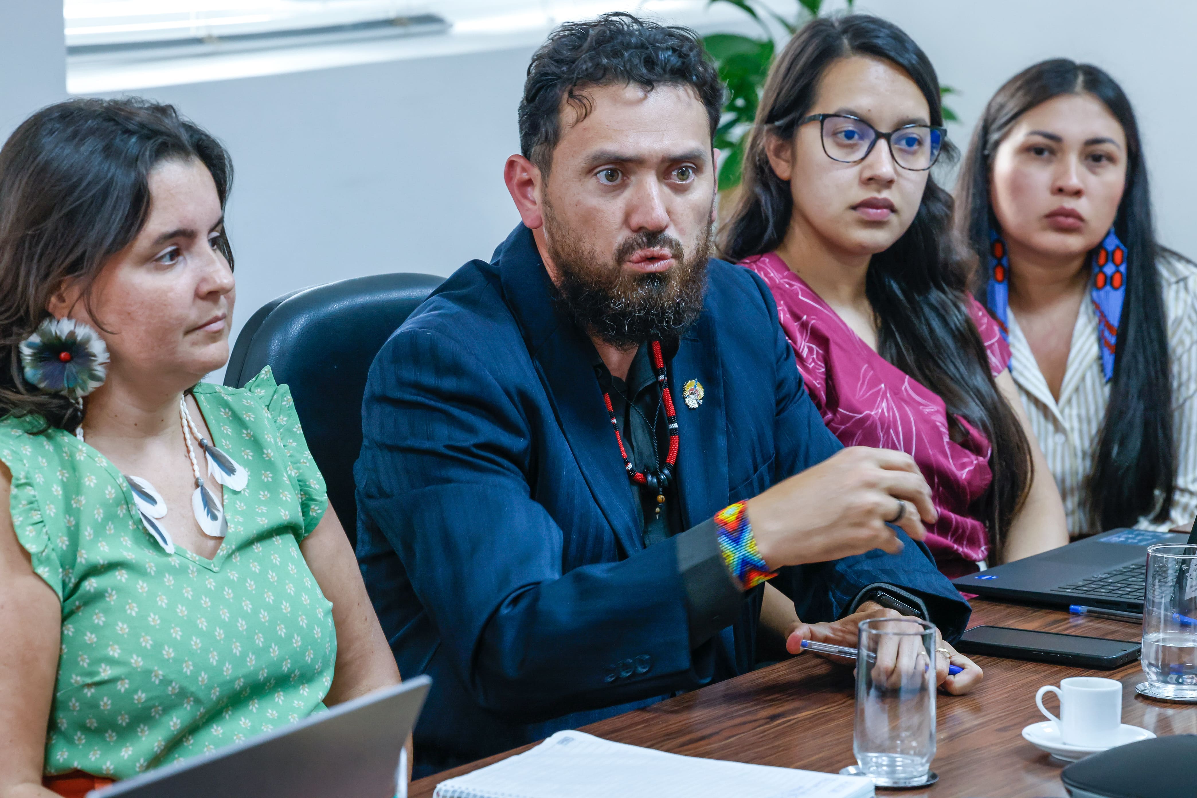 Homem de barba e paletó fala em uma reunião, ladeado por três mulheres atentas.
