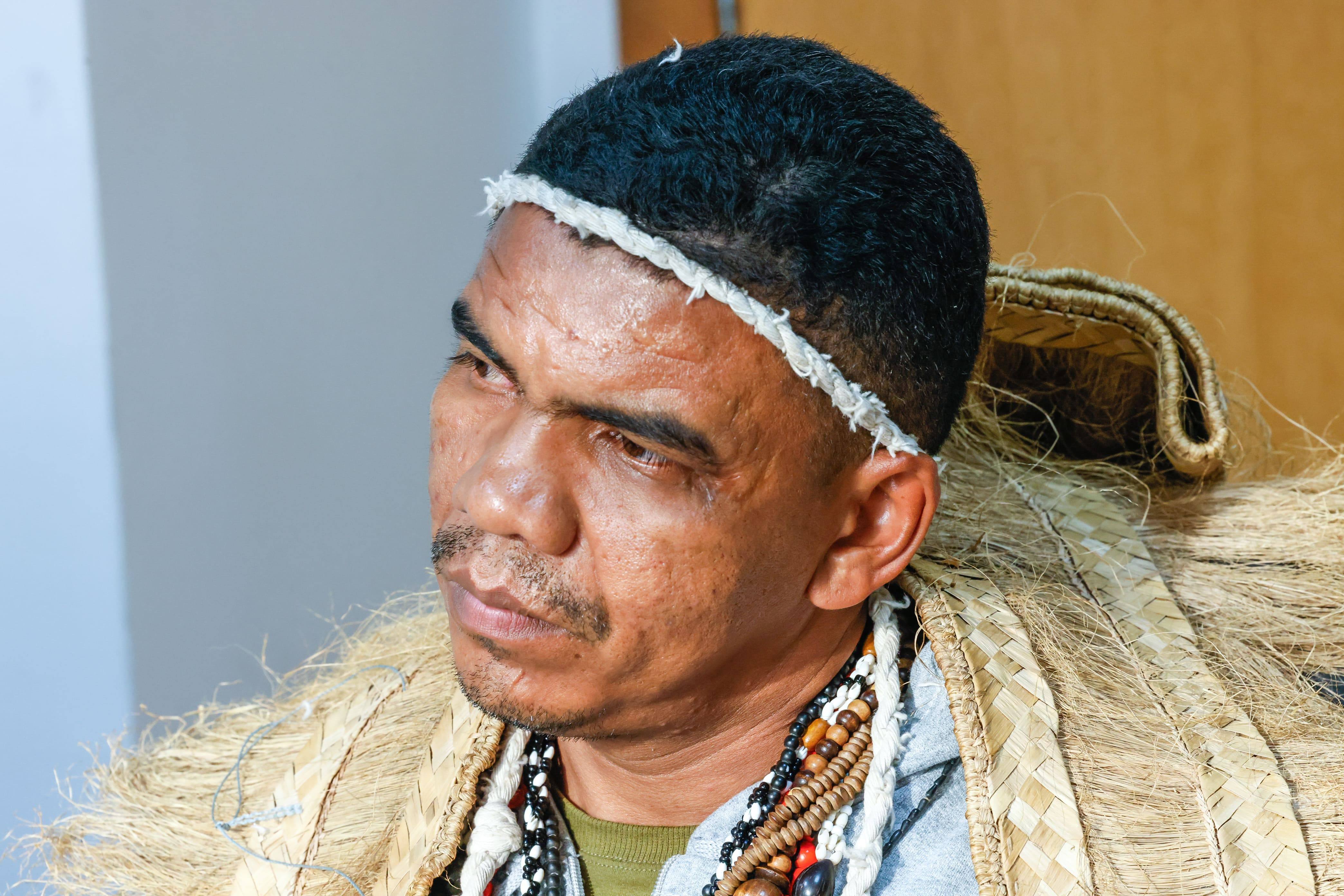 Close de um homem indígena com faixa na cabeça, colares artesanais e vestimenta de fibra natural.