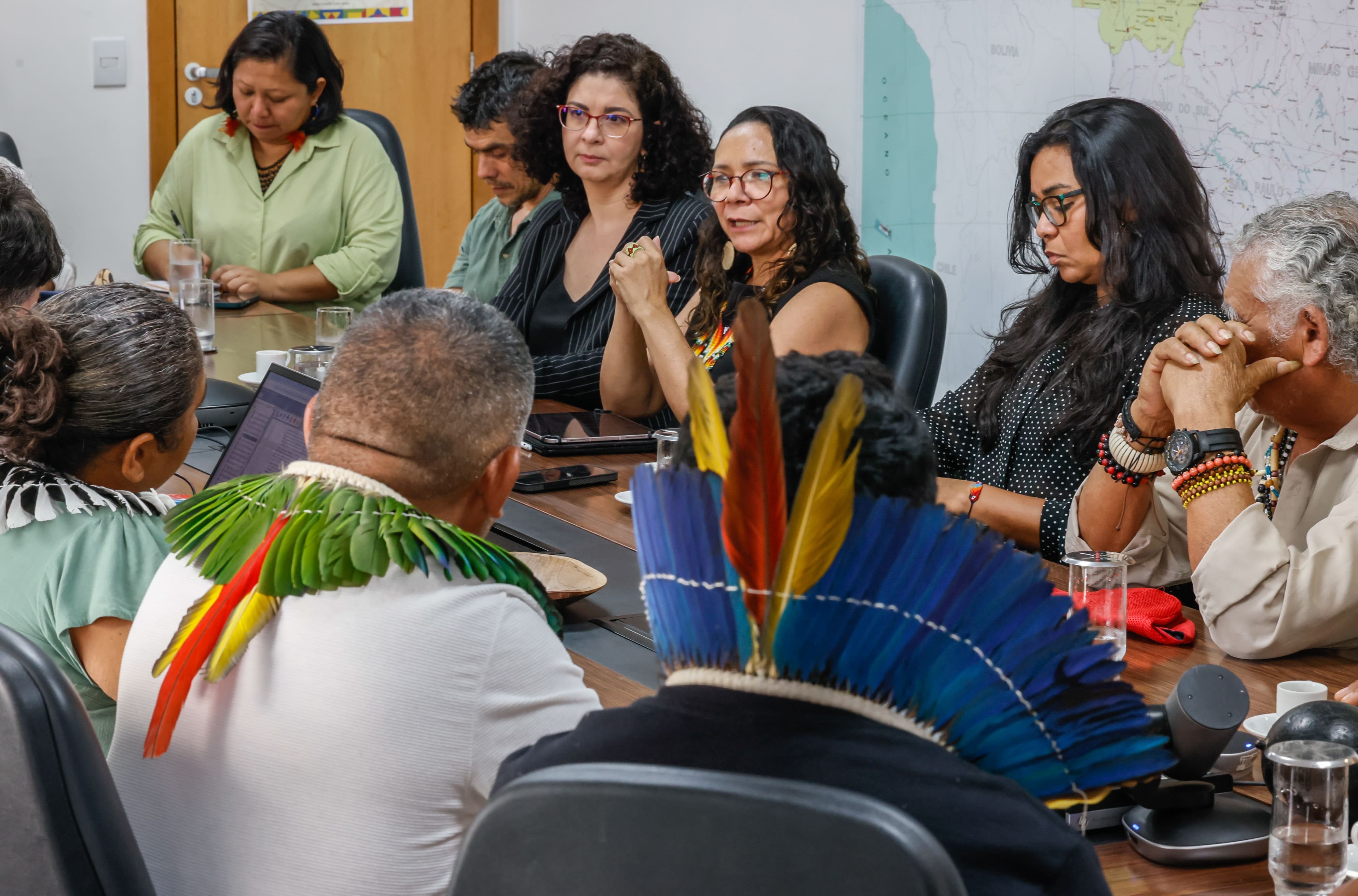 Pessoas reunidas ao redor de uma mesa de escritório, com destaque para lideranças indígenas de costas usando cocares coloridos..