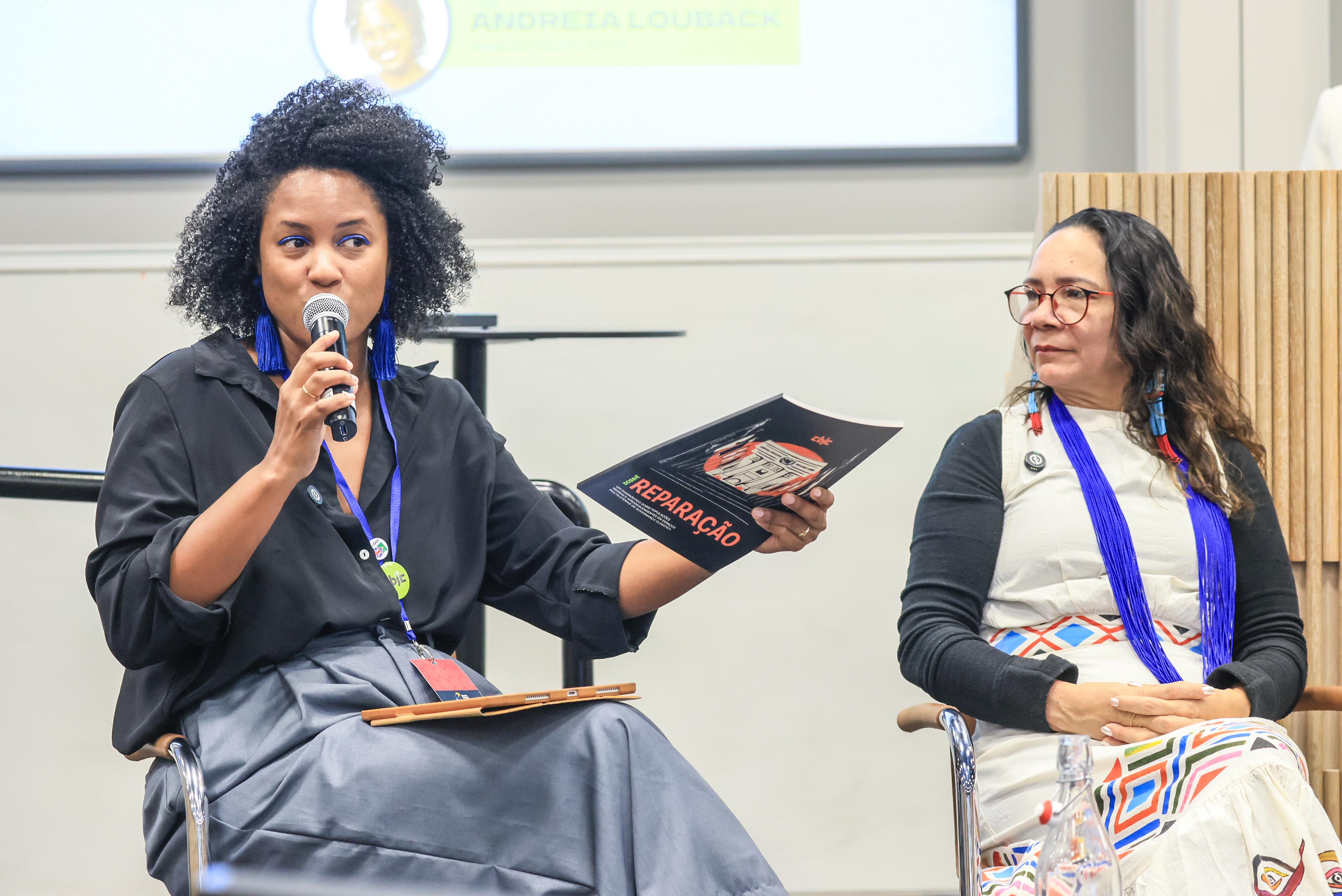 Mulher negra falando em evento, presidenta Lucia ao lado