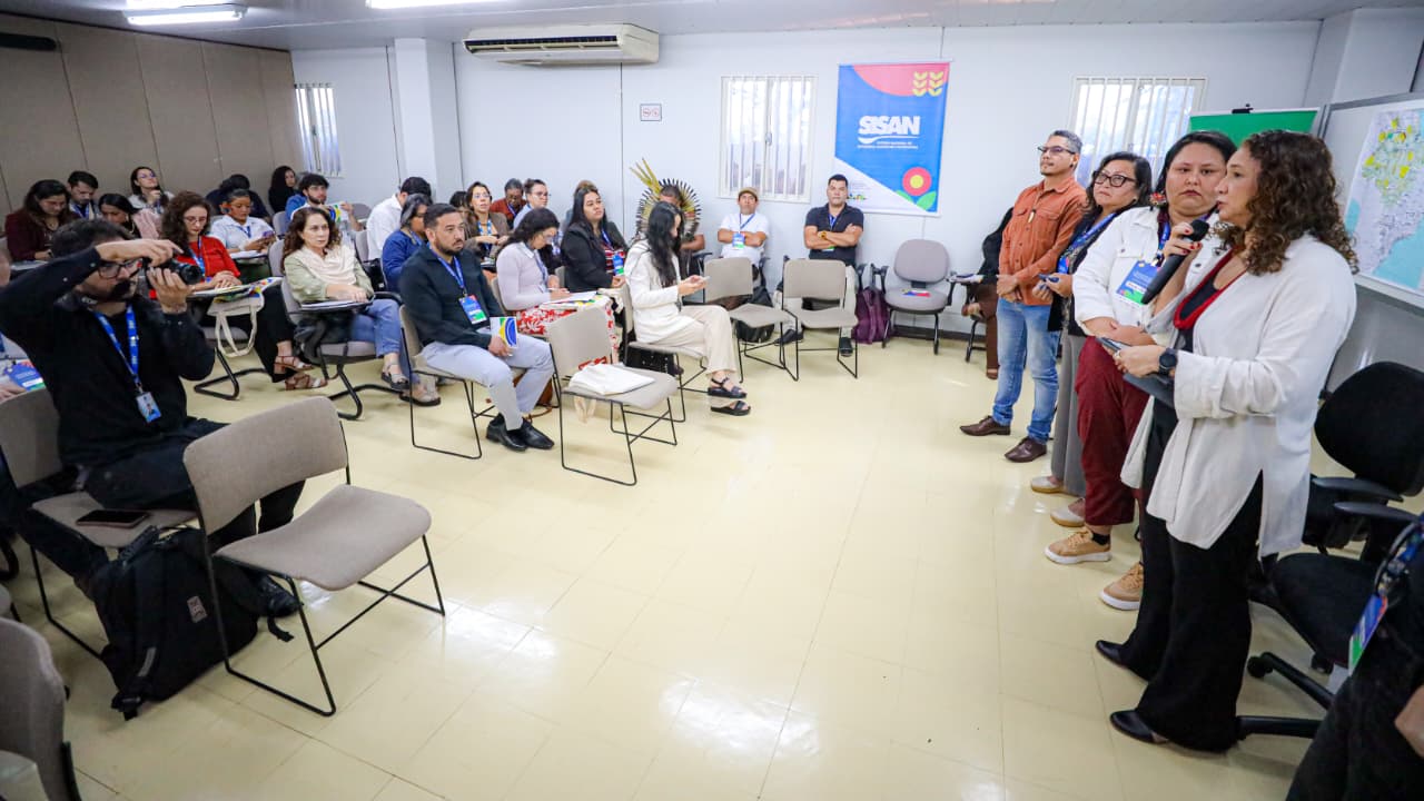 Várias pessoas sentadas em uma sala, uma mulher a frente falando