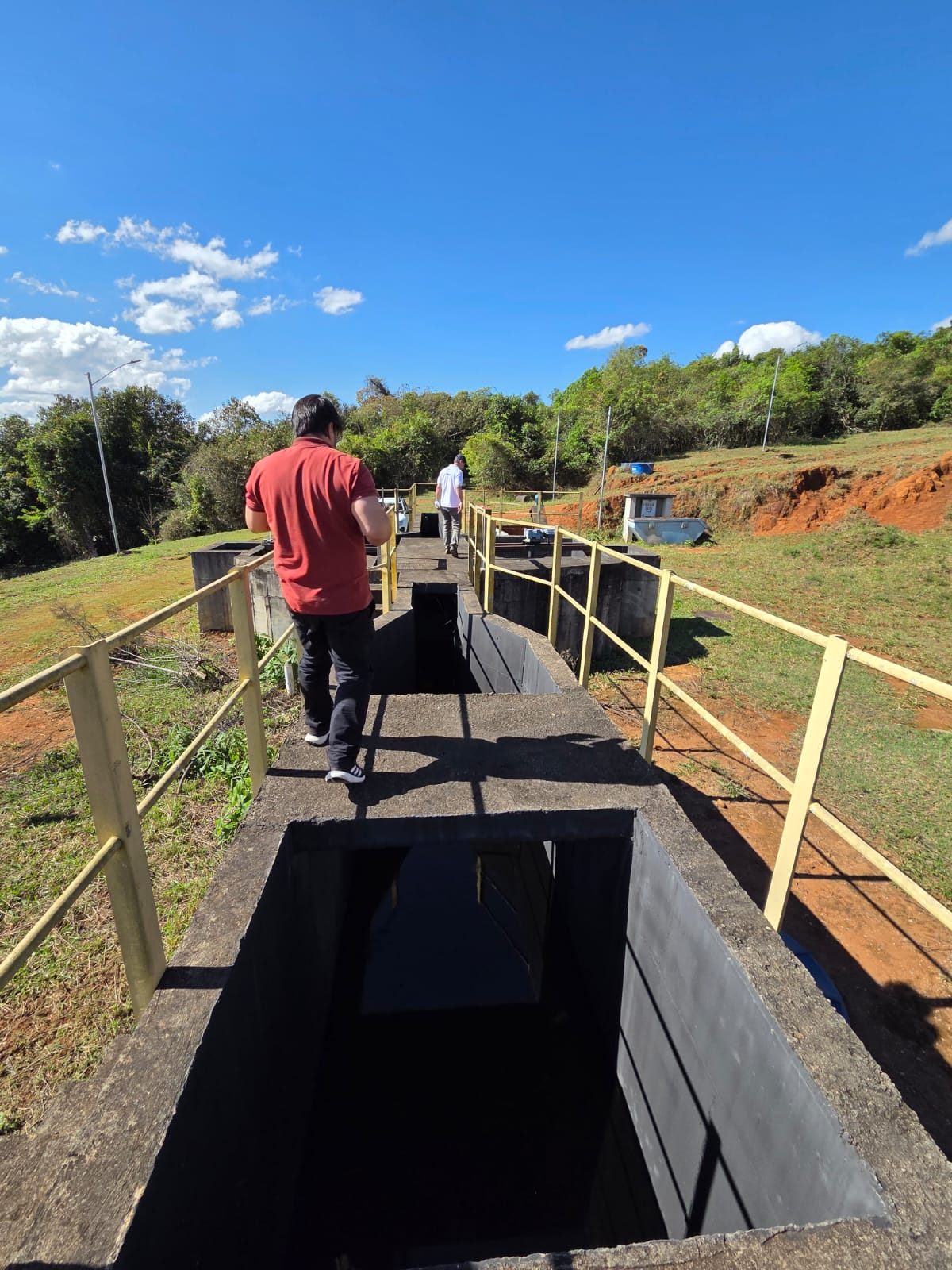 Pessoas caminham sobre passarela de concreto em estrutura para tratamento de esgoto ao ar livre.