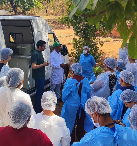 Grupo de pessoas de avental e touca ao redor de uma unidade móvel de análise em área aberta.