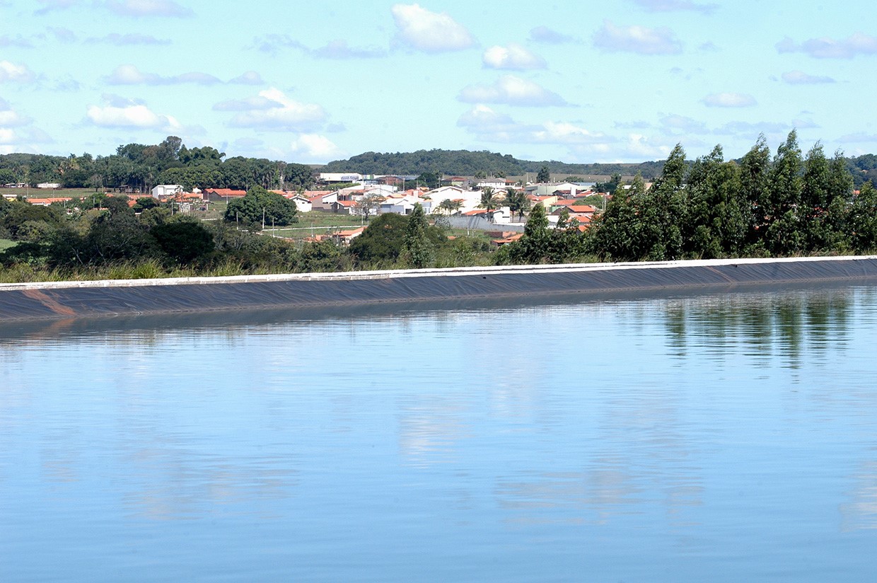 Lagoa de tratamento com água calma e casas ao fundo sob céu azul com poucas nuvens.