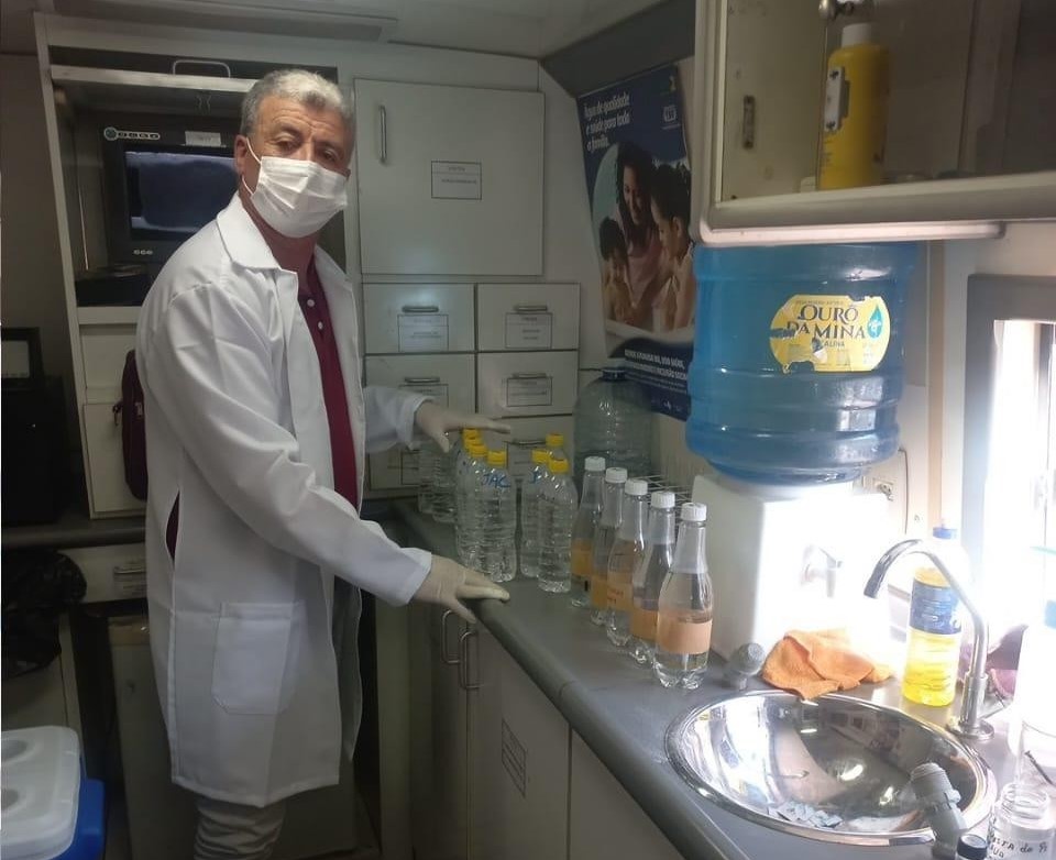 Homem de jaleco e máscara organizando garrafas de vidro com amostras de água em laboratório.