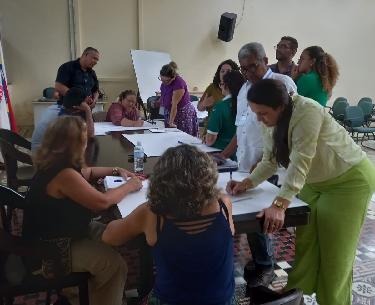 Pessoas reunidas em volta de uma mesa grande escrevendo e analisando folhas de papel.