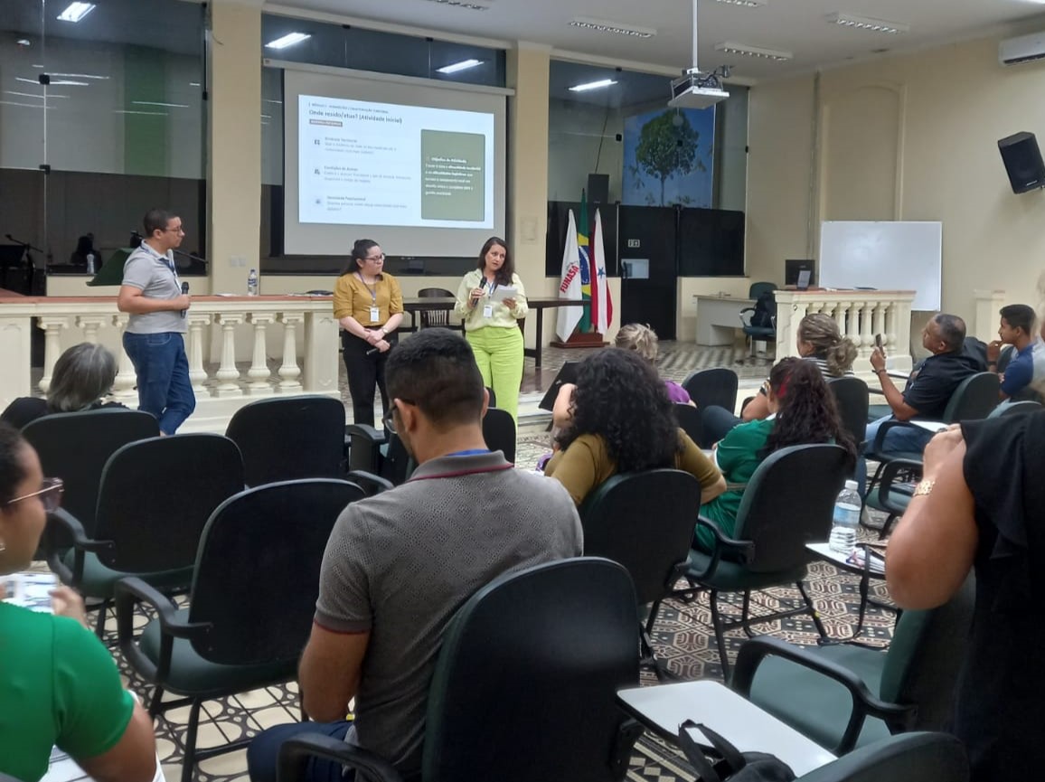 Palestrantes em pé apresentando slide para uma plateia sentada em auditório.