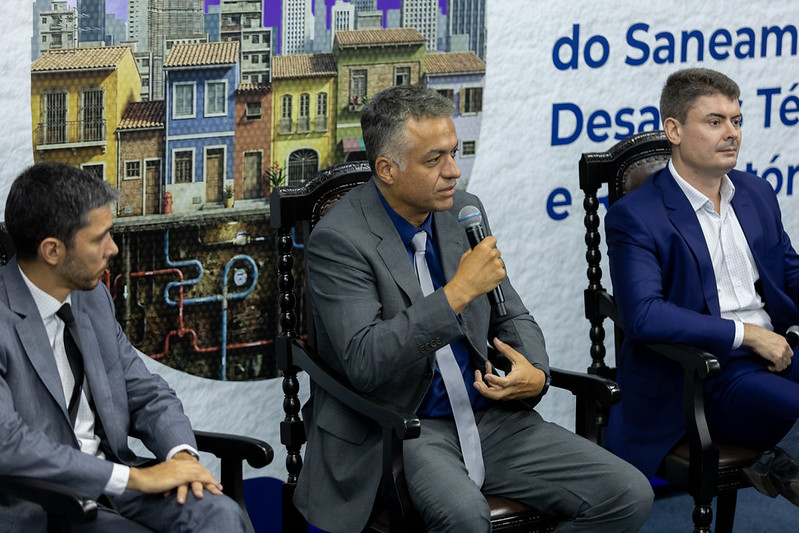 Homem de terno fala ao microfone sentado ao lado de outros palestrantes em evento sobre saneamento.