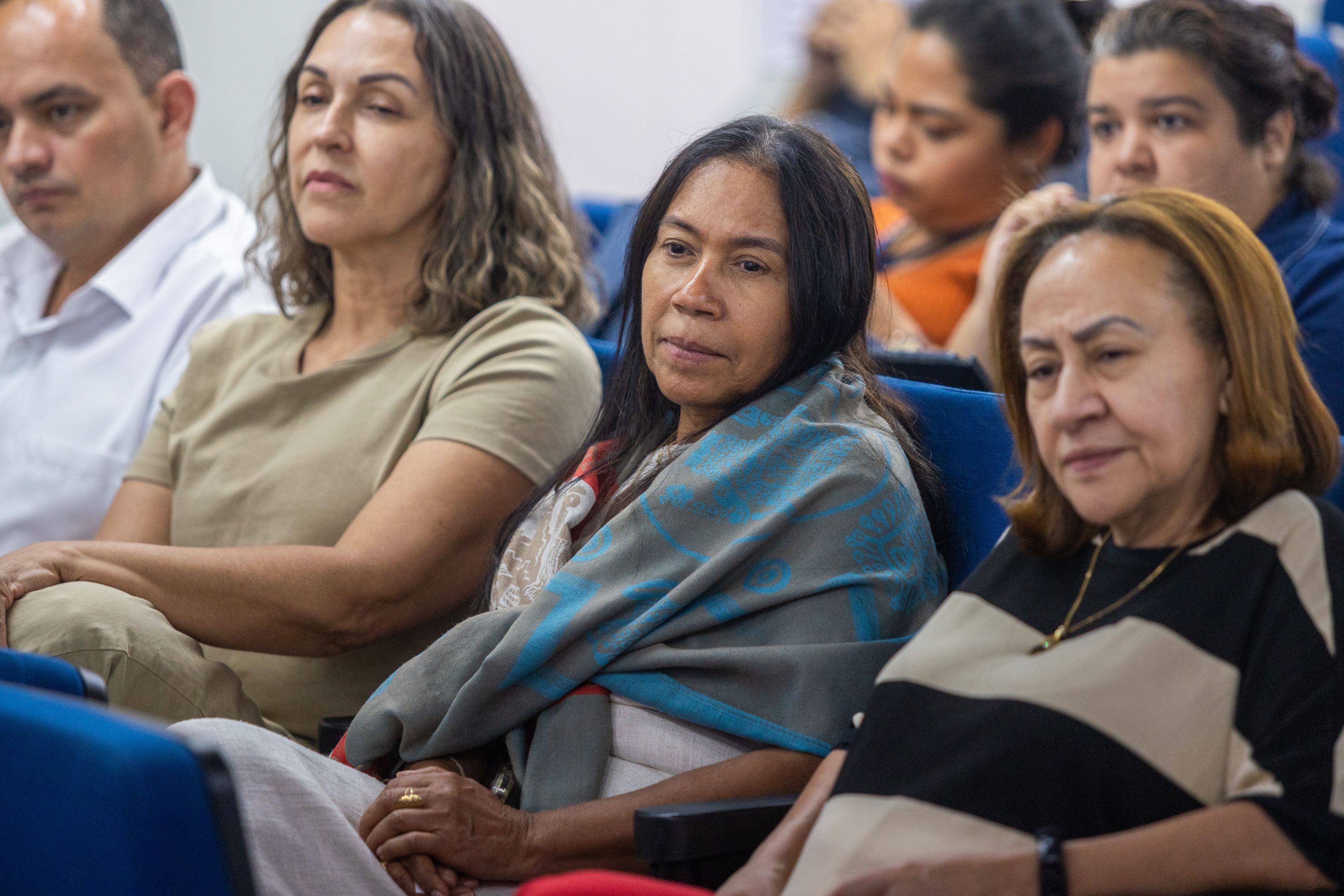 Pessoas, em sua maioria mulheres, sentadas em cadeiras azuis atentas a uma apresentação.