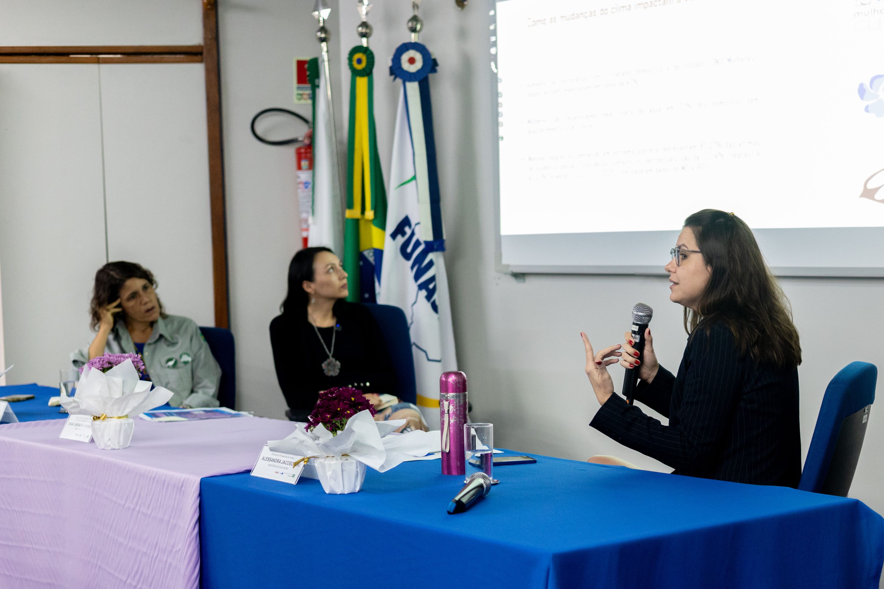 Mulher de óculos fala ao microfone em mesa de debate, com duas ouvintes e projeção ao fundo.