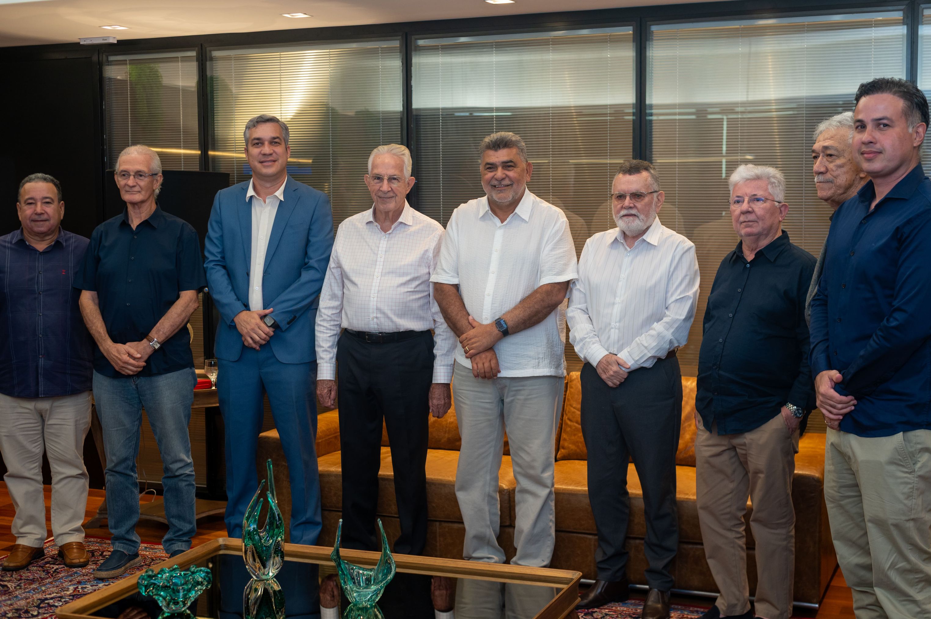 Nove homens em trajes esporte fino posam lado a lado em sala de reuniões.