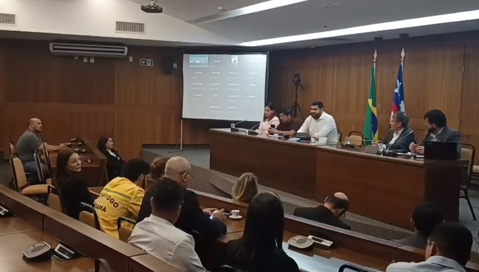 Autoridades compostas por homens e mulheres sentadas à mesa de reunião diante de um auditório.