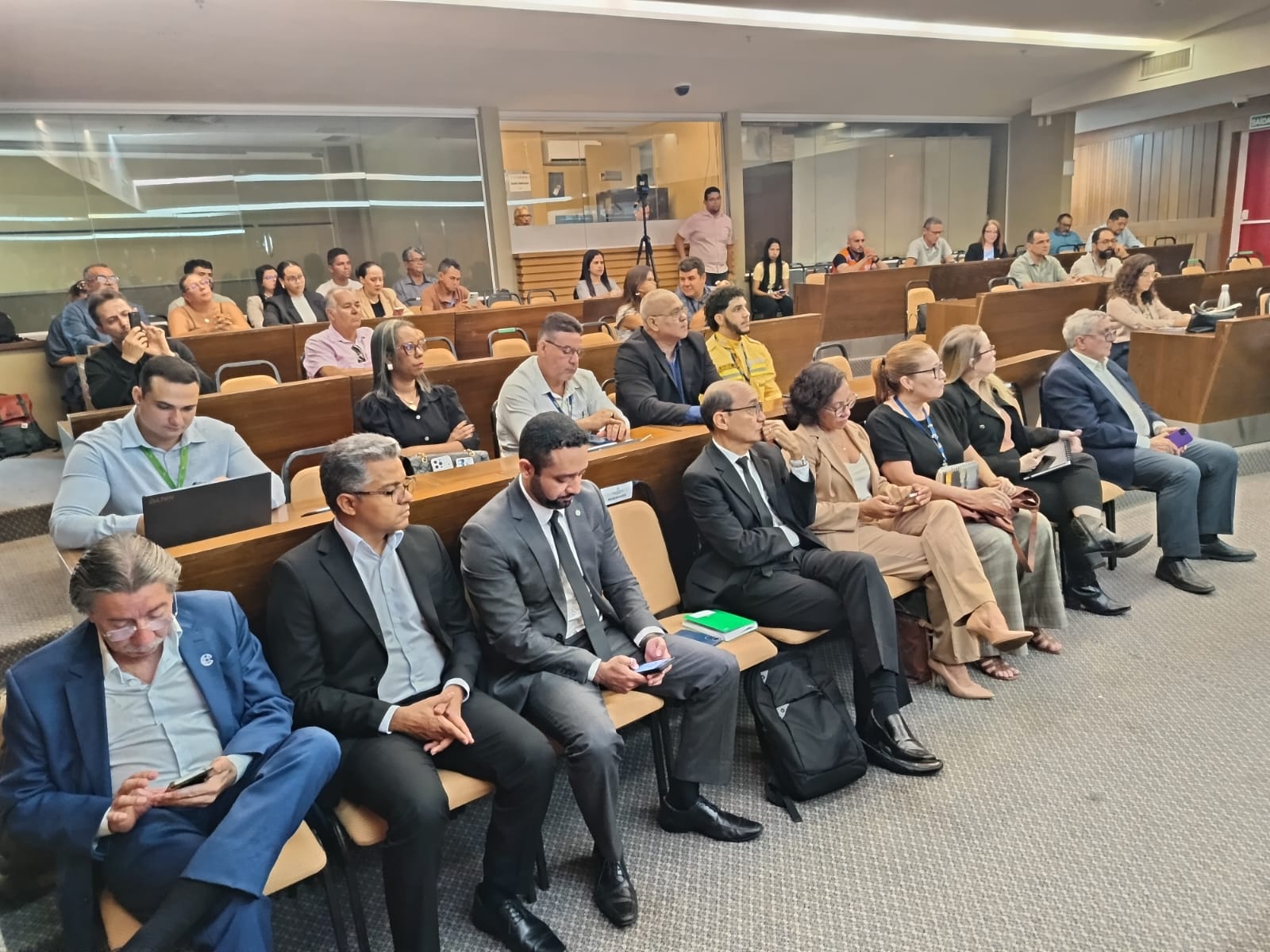 Pessoas vestidas formalmente sentadas nas bancadas de um auditório acompanhando reunião.