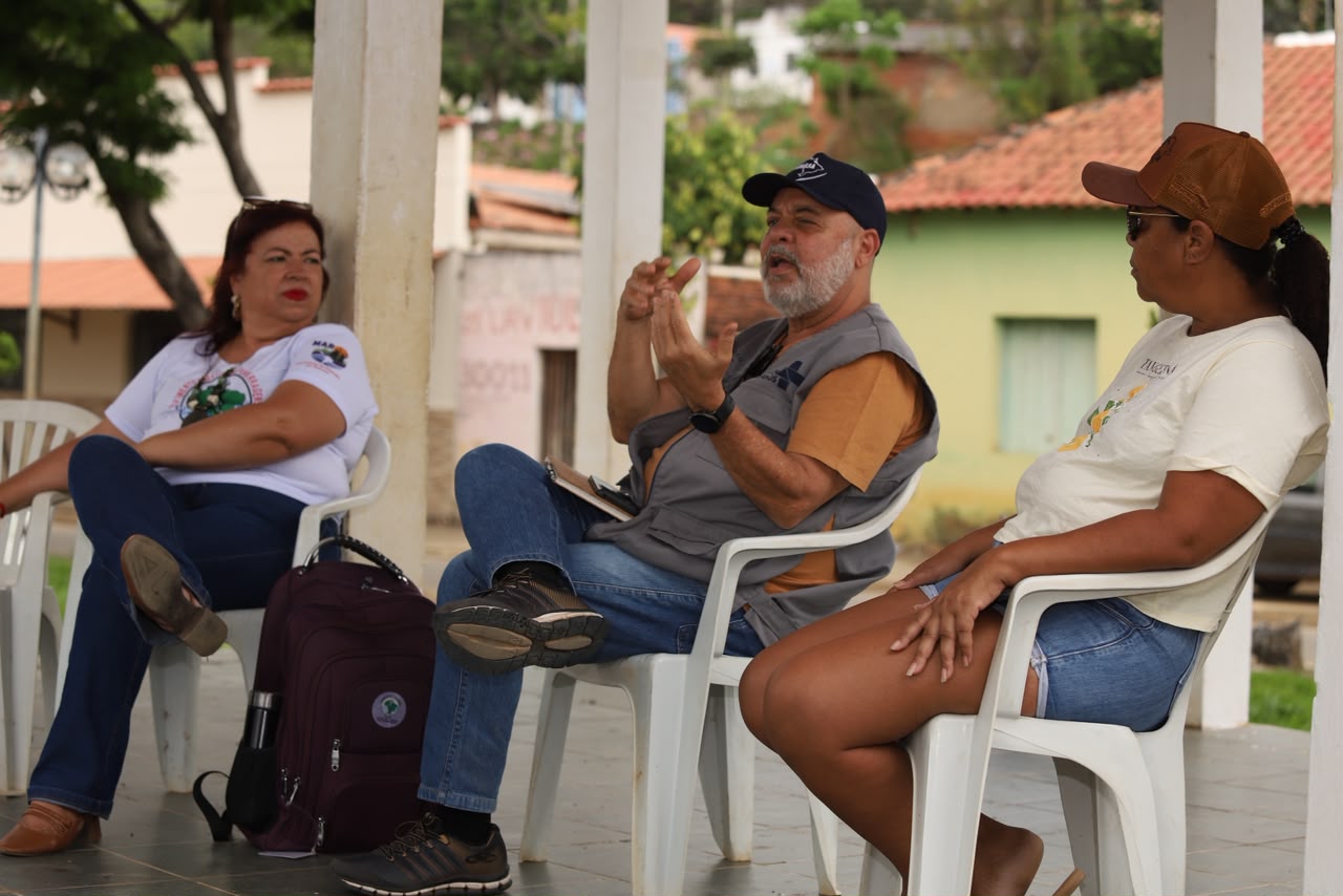 Homem de boné e colete gesticula enquanto fala sentado em roda de conversa com duas mulheres.