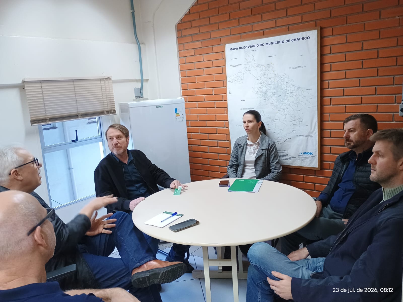 Pessoas reunidas em torno de uma mesa redonda em sala com parede de tijolos e mapa do município.