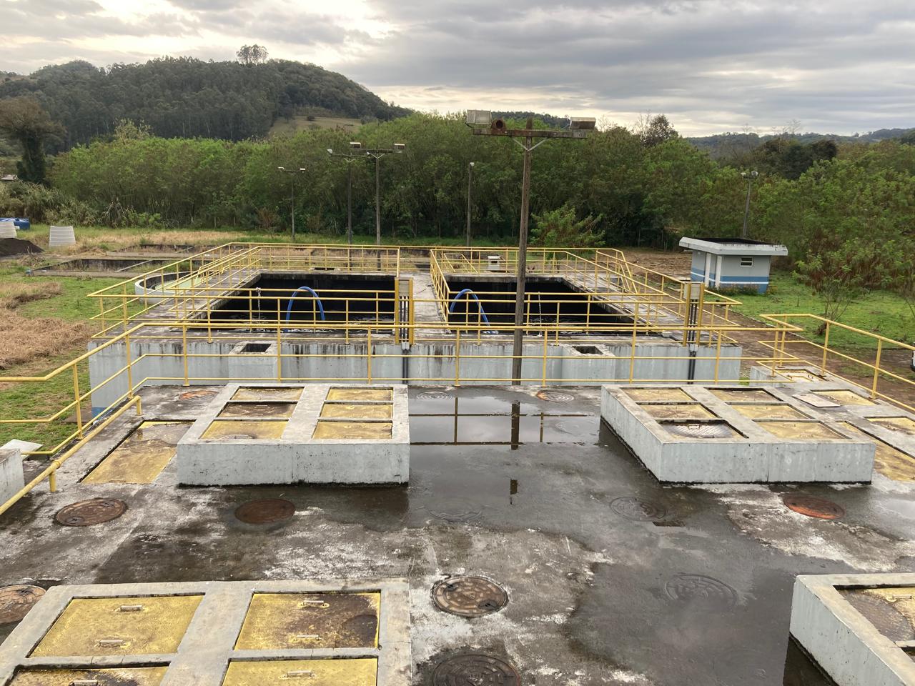 Estação de tratamento de esgoto com tanques de concreto, grades amarelas e vegetação ao fundo.