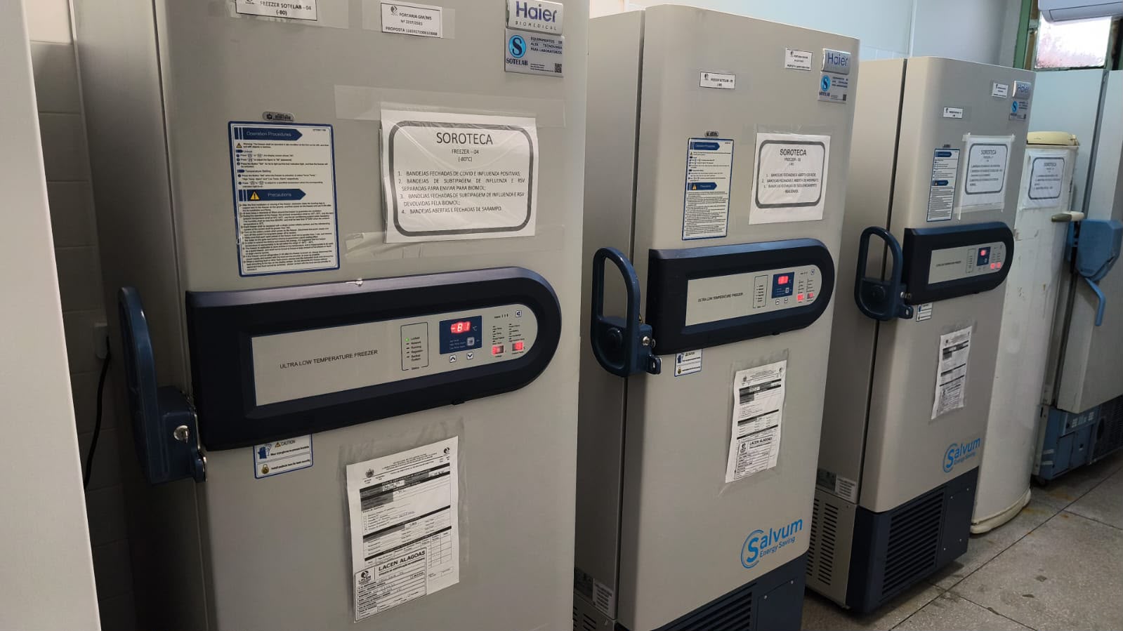 Três ultrafreezers verticais de laboratório alinhados, com papéis informativos colados nas portas.