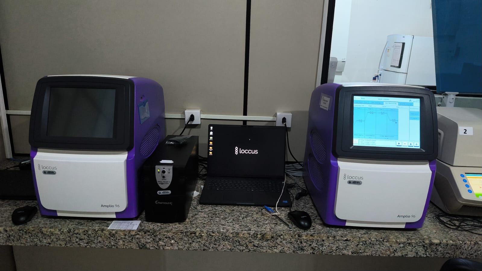 Dois aparelhos de PCR roxos e brancos ao lado de um notebook sobre bancada de laboratório.