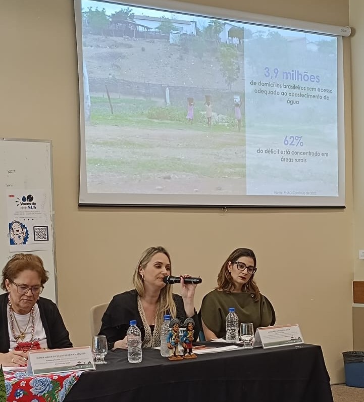 Mulheres em mesa de debate sob projeção de dados sobre falta de acesso à água no Brasil.
